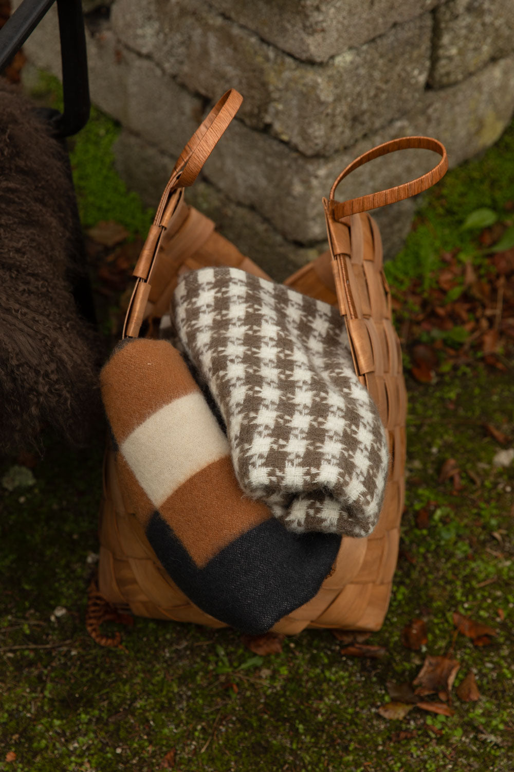 Woven basket with leather handles holding a brown and cream houndstooth lambswool blanket and an orange block-check blanket on autumn grass