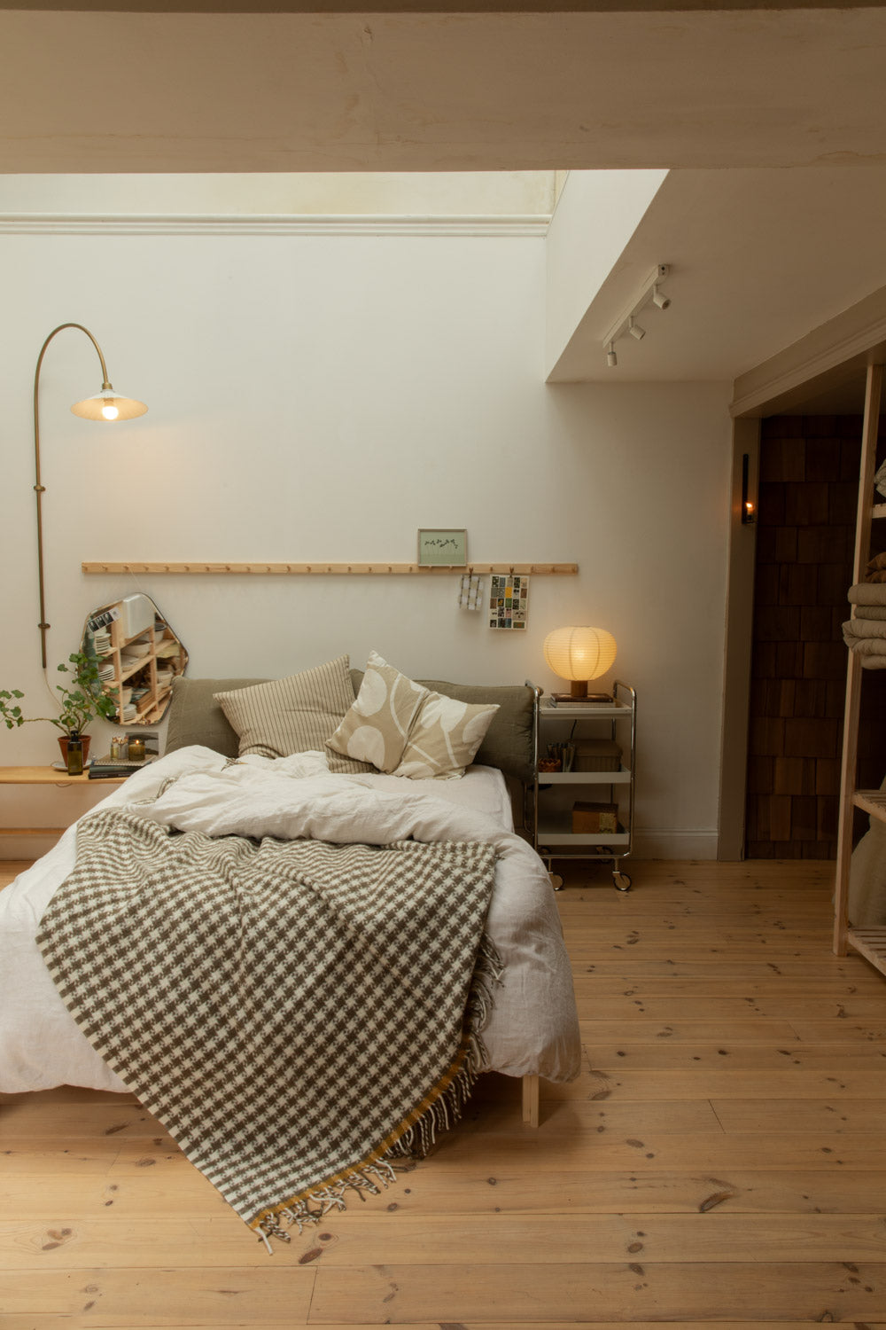 Calm bedroom scene with taupe houndstooth wool blanket on white linen bed, Blomus Kami table lamp, brass floor lamp and pine peg rail.