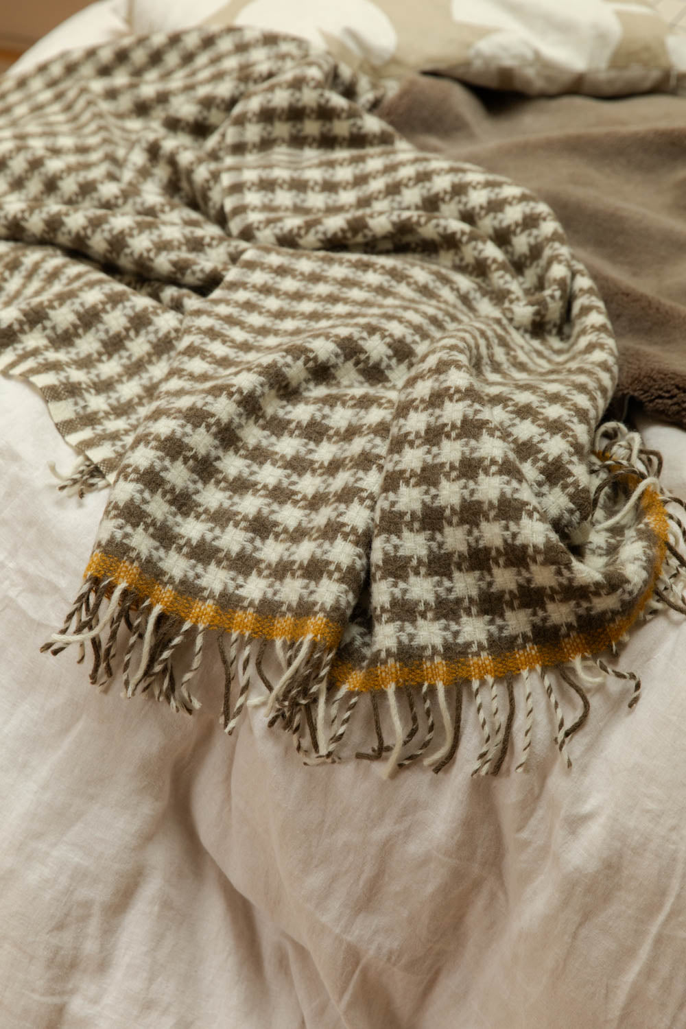 Close-up of houndstooth wool throw with mustard stripe and twisted fringe detail draped on white linen bed.