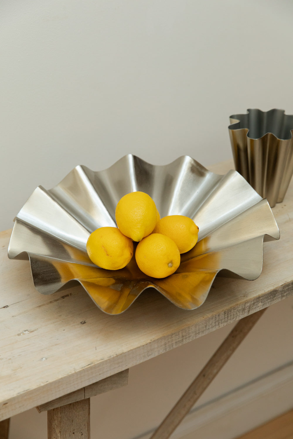 Angled close-up of House Doctor Flood display bowl in brushed silver filled with lemons with matching ribbed silver vase in background.