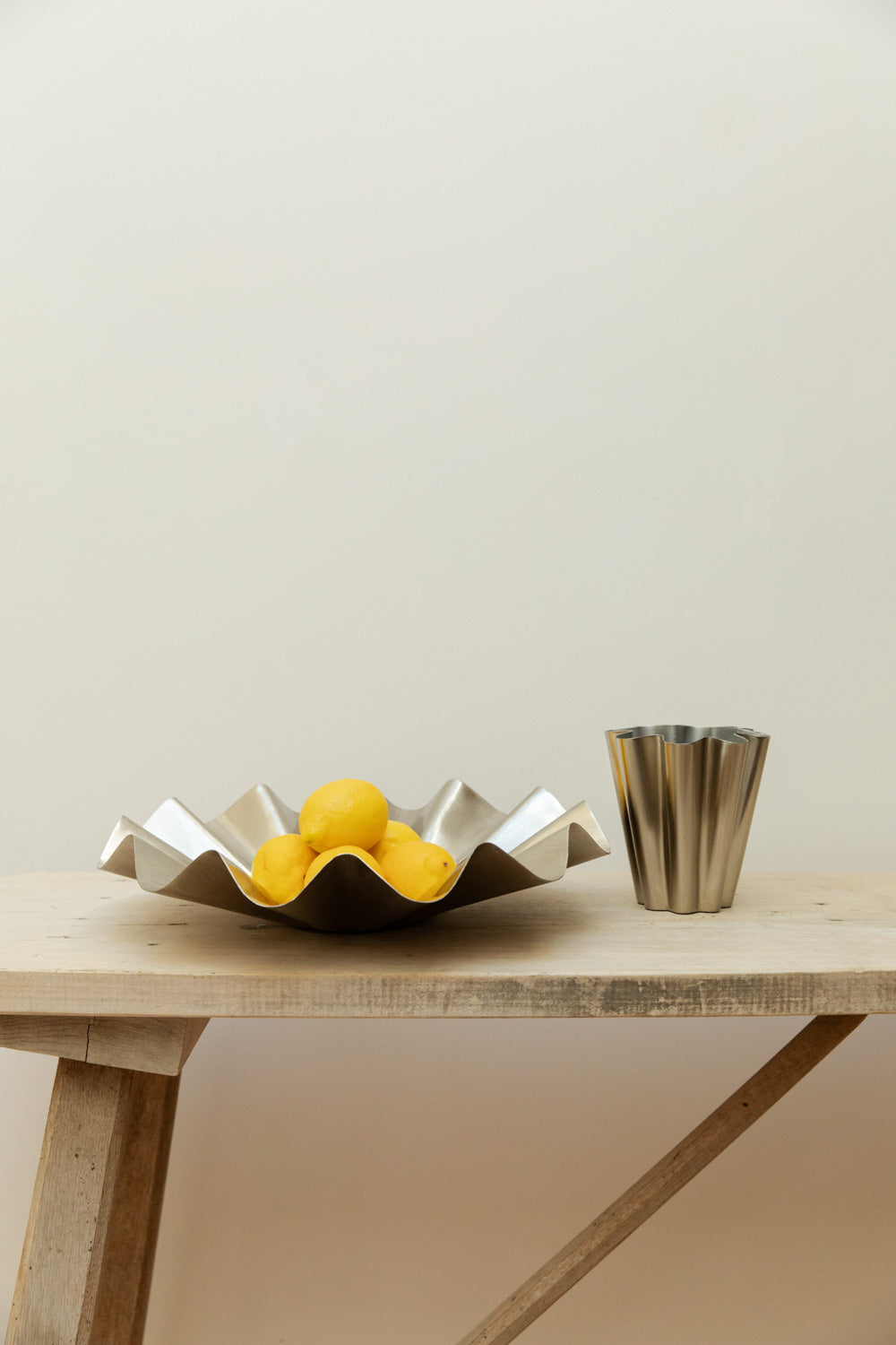 House Doctor Flood display bowl in brushed silver with lemons styled alongside matching ribbed silver vase on a rustic pale wood table.