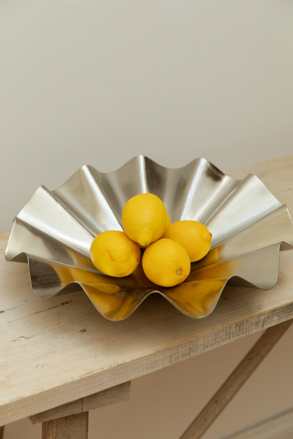 Close-up of House Doctor Flood display bowl in brushed silver filled with lemons on a rustic pale wood table.