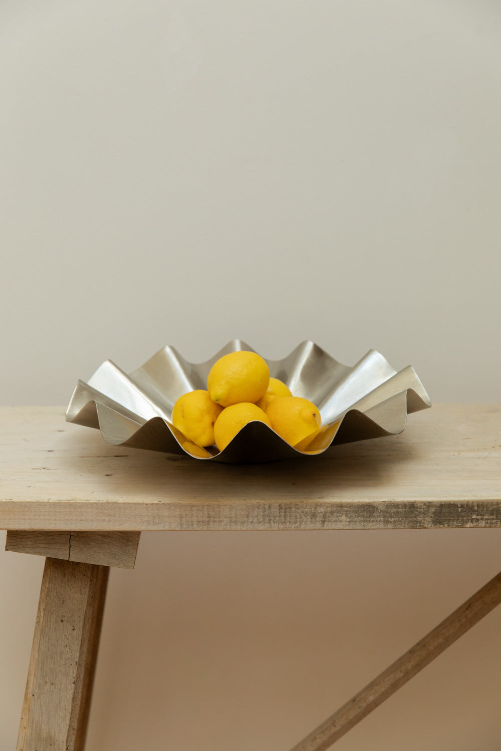 House Doctor Flood display bowl in brushed silver holding lemons on a rustic pale wood table, side view.