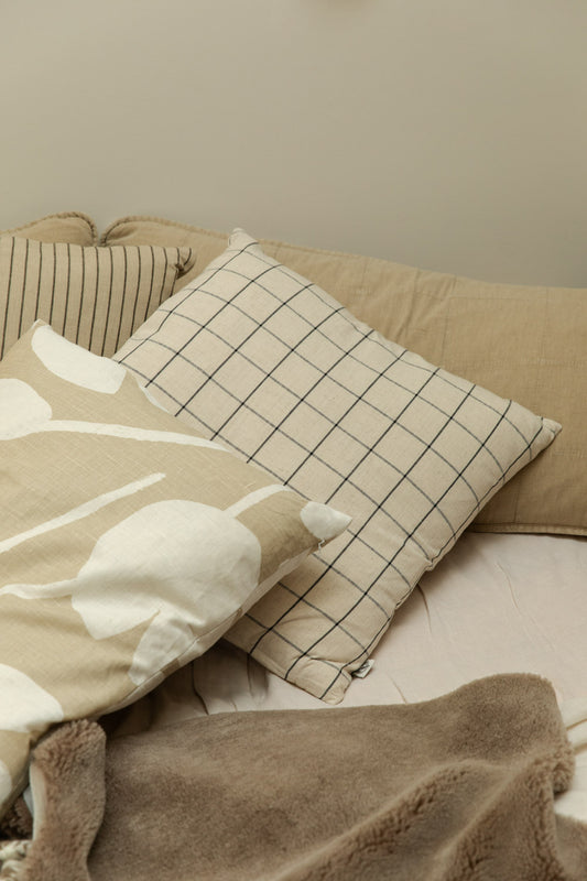 House Doctor Join grid check linen cushion cover and Water Lilies sand cushion cover styled together on a bed with a pinstripe cushion and taupe lambswool rug.