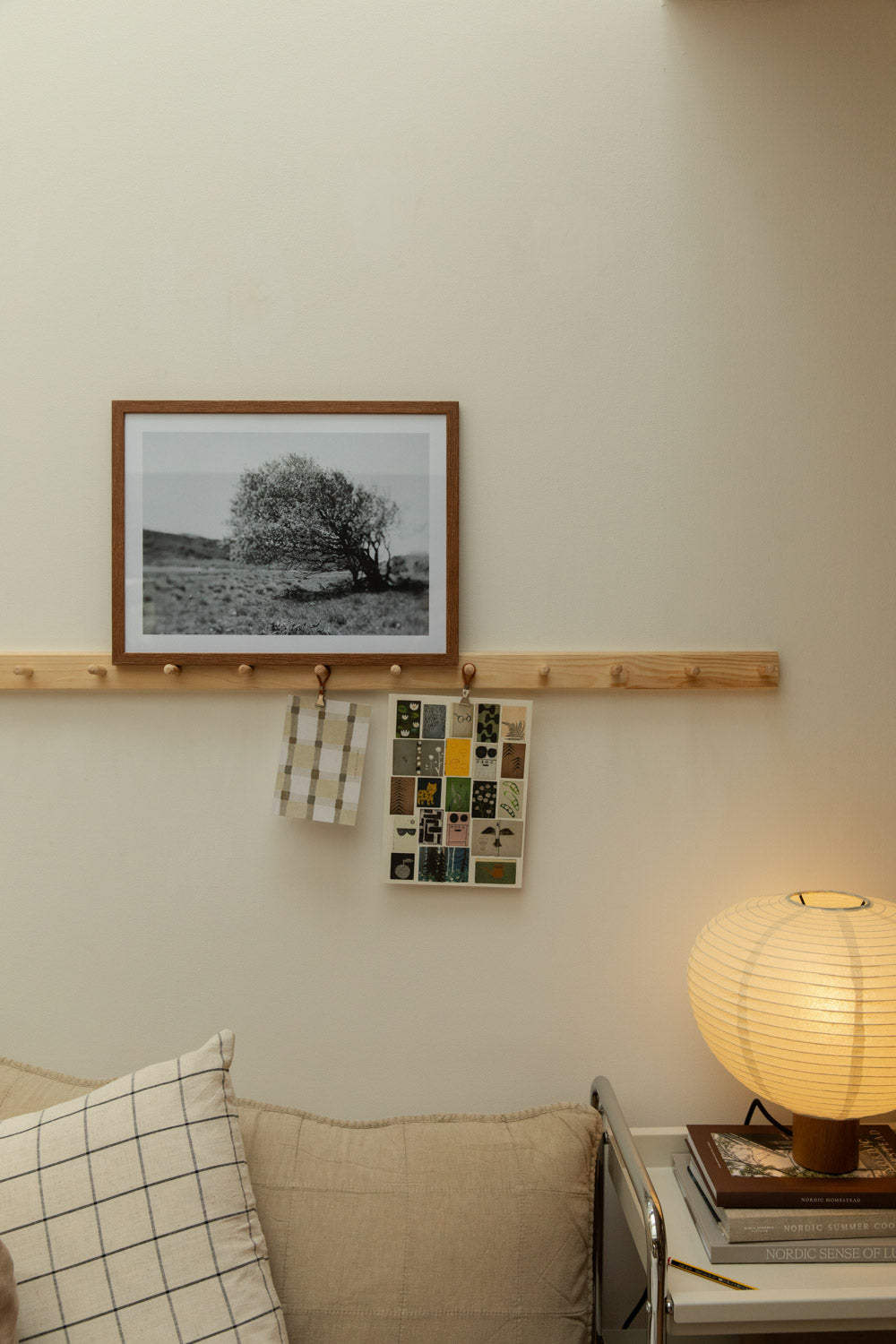 House Doctor Join grid check linen cushion cover on a sofa below a shaker peg rail styled with a black and white tree art print in a walnut frame, hanging cards and a glowing paper lantern table lamp.