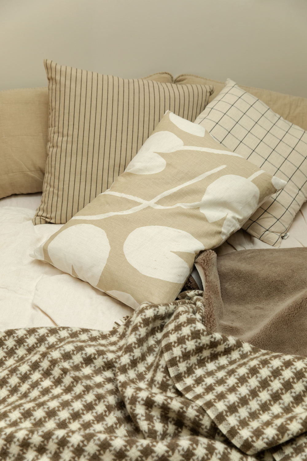 Fine Little Day Water Lilies sand cushion, Kare striped cushion and Join cushion against a fog linen headboard cushion on a bed with Klippan Houndstooth blanket and a taupe sheepskin rug.