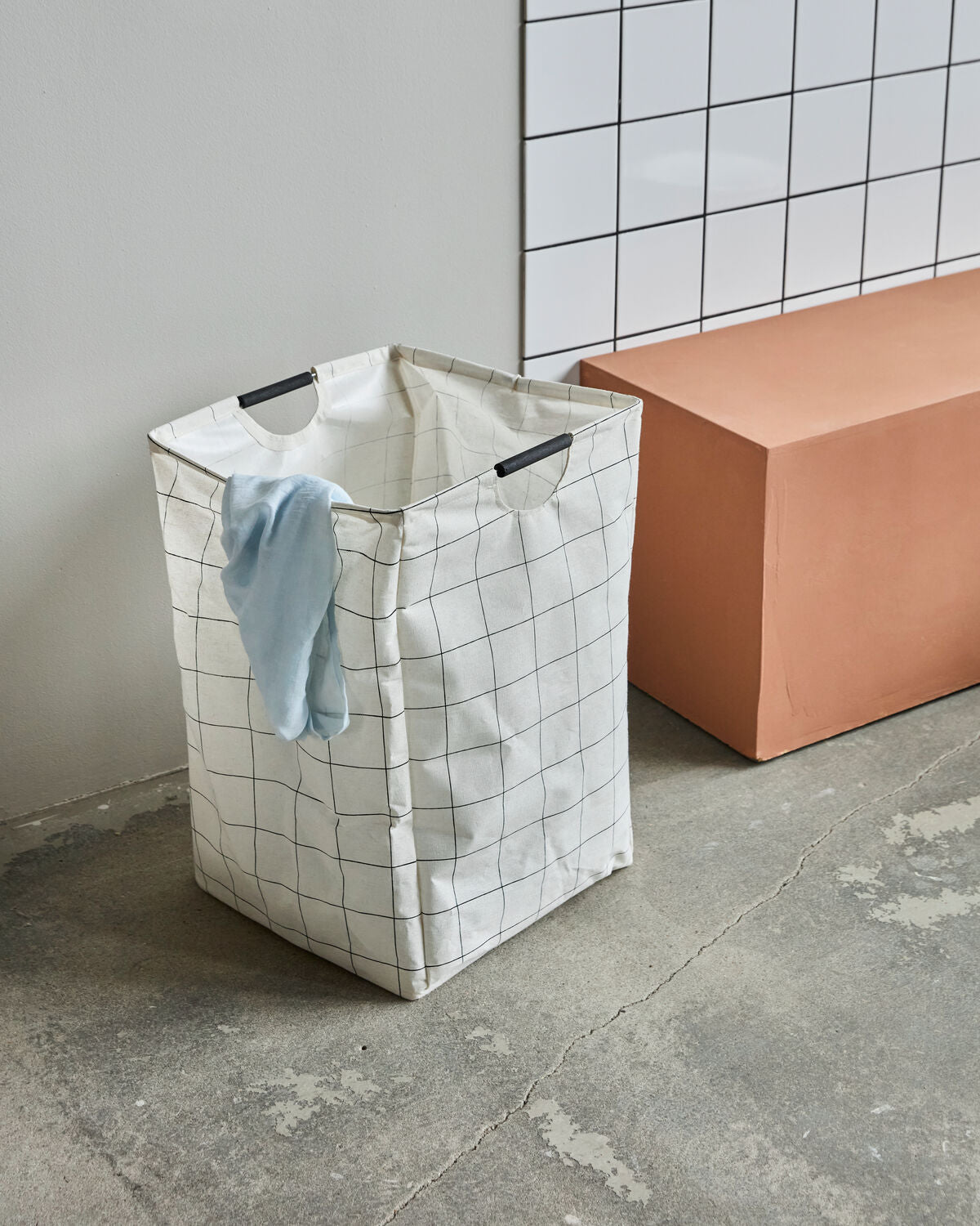 House Doctor HD Squares laundry bag standing open with a blue cloth coming out the top on a concrete floor with white tiles behind a a terracotta block next to it. 
