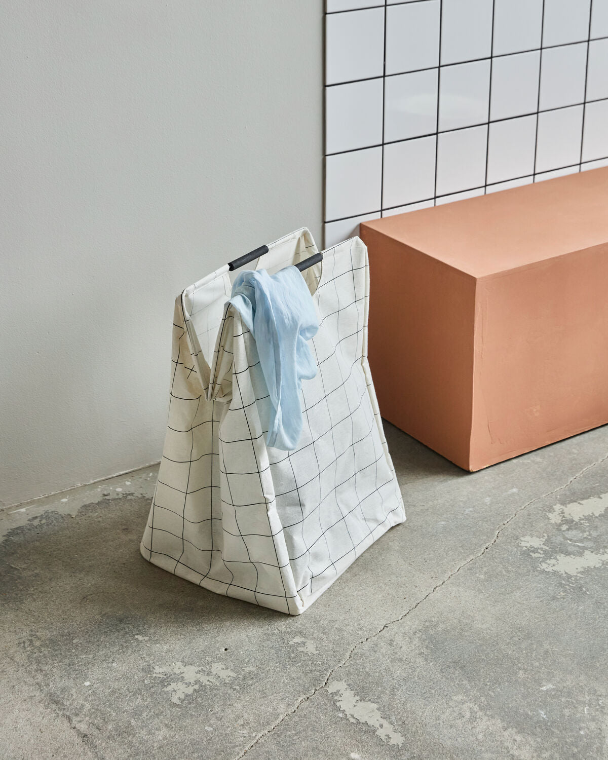 House Doctor HD Squares laundry bag standing closed with a blue cloth coming out the top on a concrete floor with white tiles behind a a terracotta block next to it. 