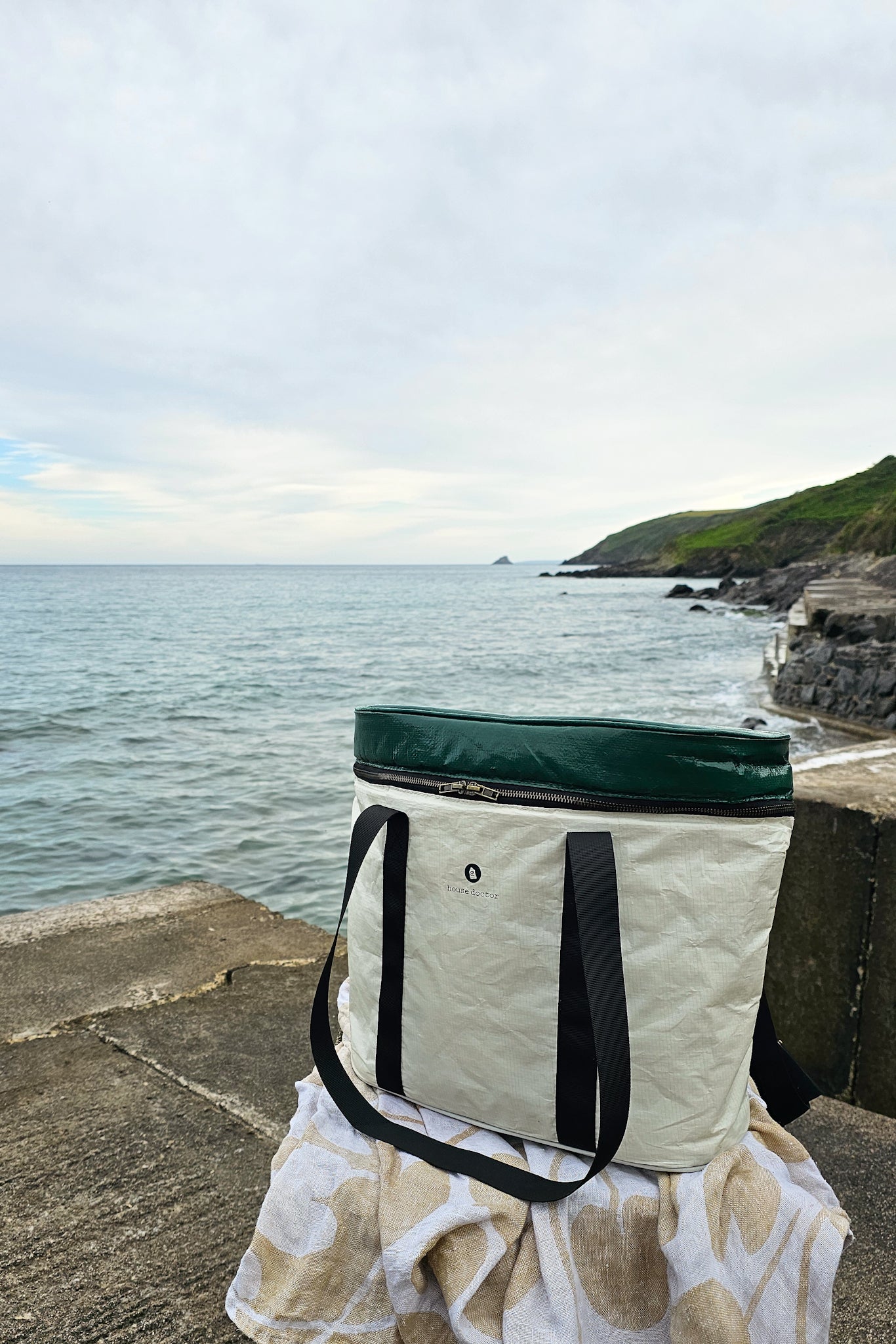 House Doctor Picnic Cooling Bag in sand and green, photographed on a Cornish harbour wall with coastal cliffs and sea in the background.