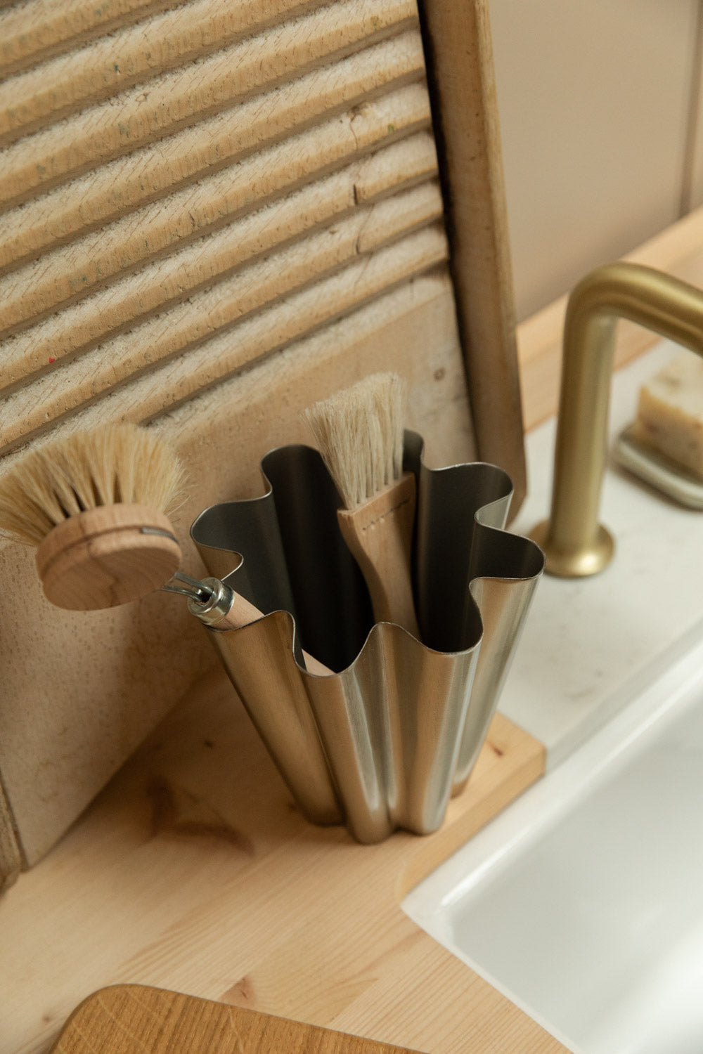 House Doctor Flood small brushed silver vase holding natural dish brushes beside a Belfast sink, vintage washboard and brass tap.
