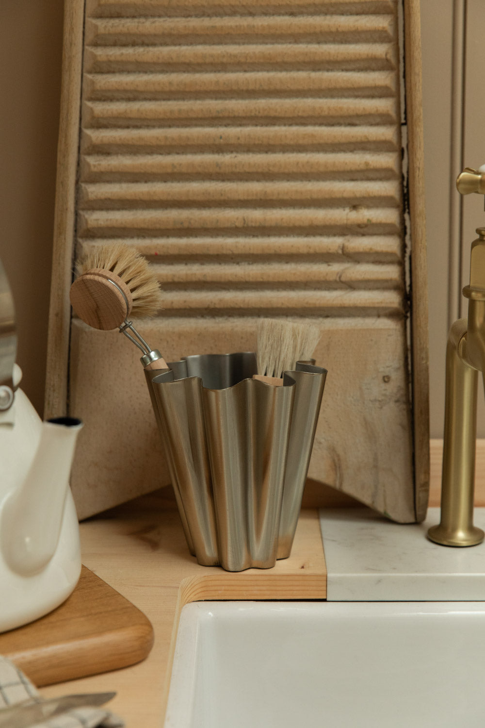 Close detail of House Doctor Flood small brushed silver vase with natural dish brushes beside a brass tap, vintage washboard and Belfast sink.