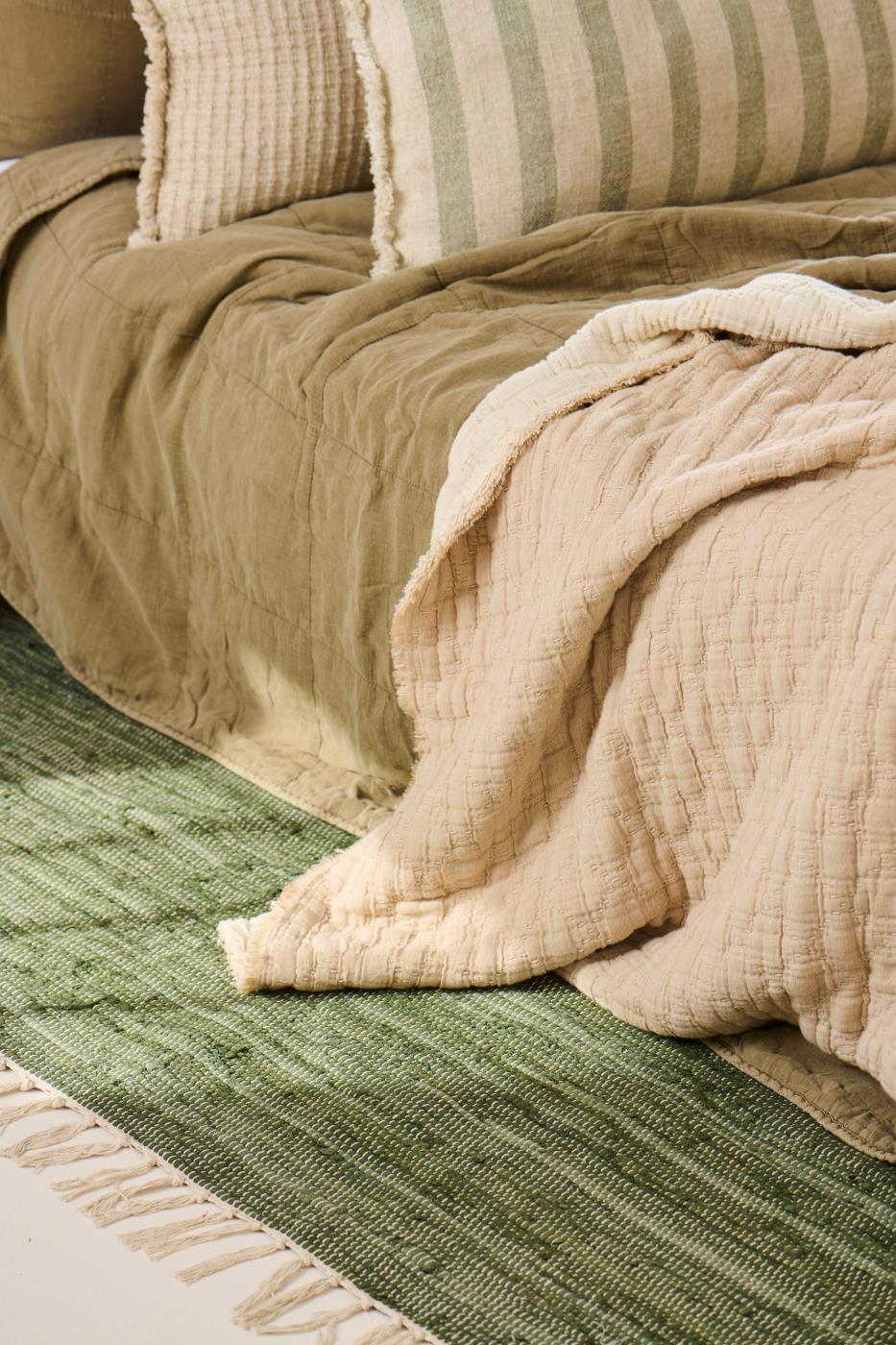 Close-up of a textured beige blanket draped over a green rug with pillows in the background.