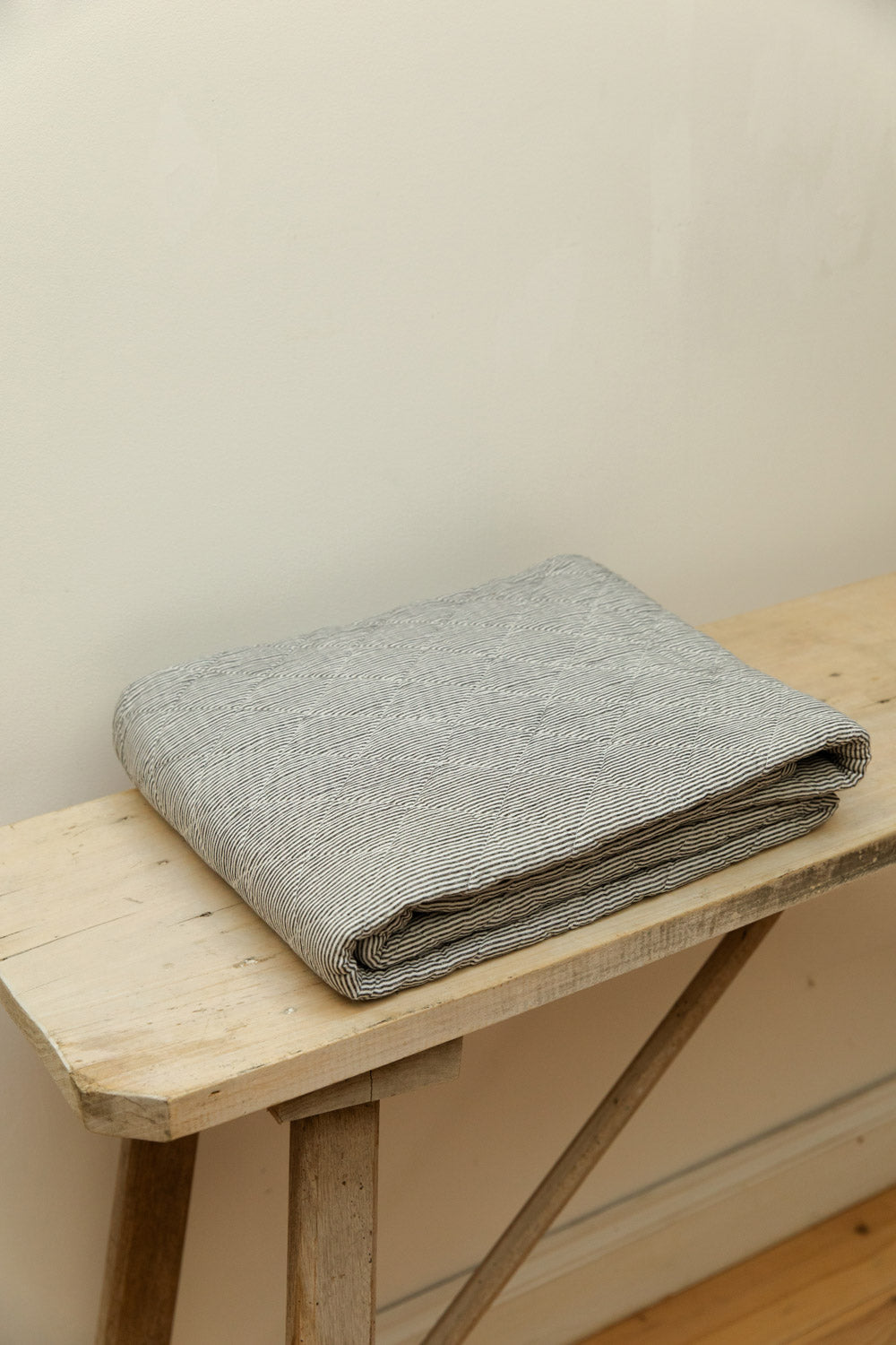 Close view of Ib Laursen grey stripe quilt neatly folded on a pale wood trestle bench, showing fine stripe and quilted texture detail.