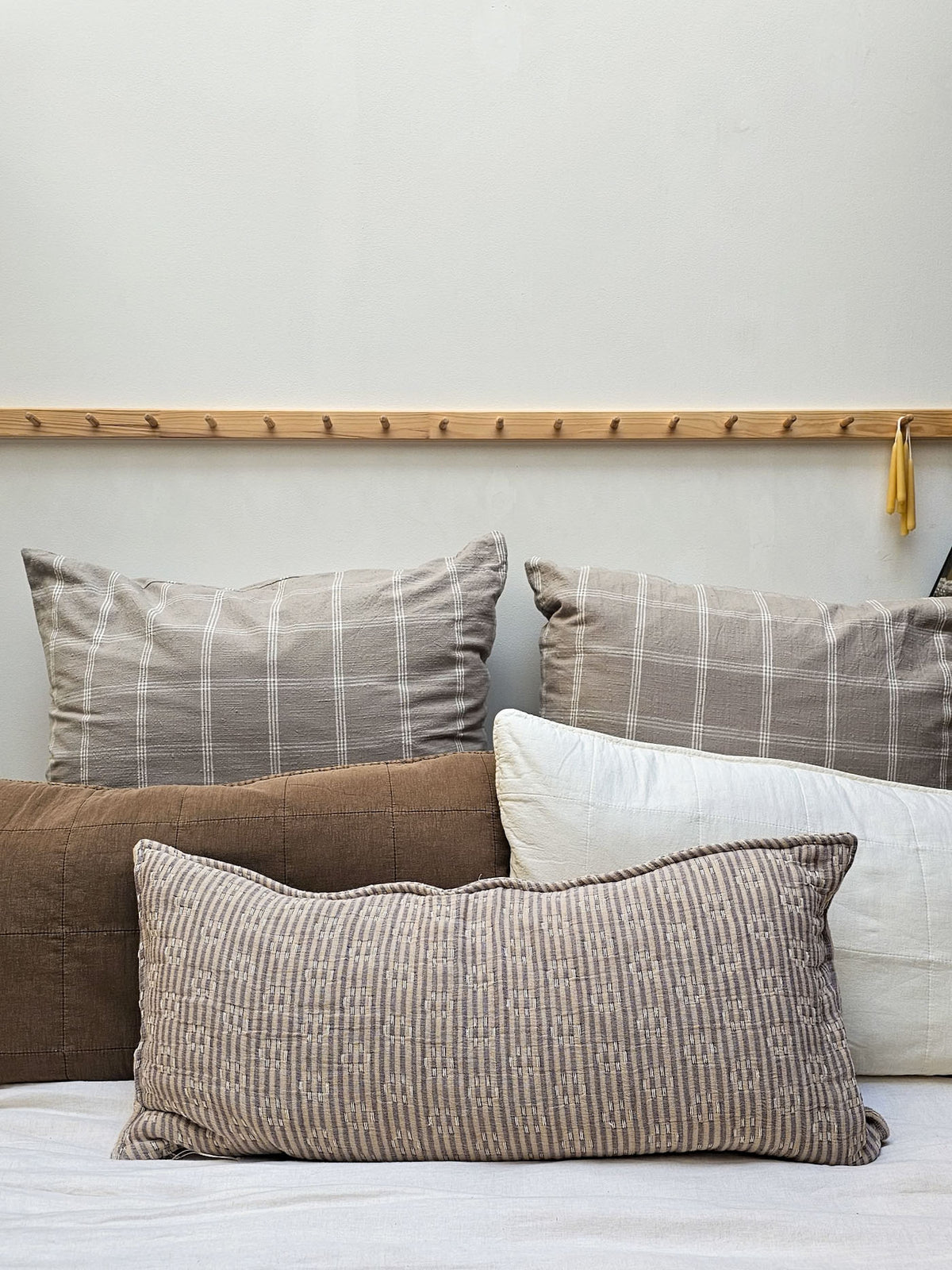 A collection of textured throw pillows in neutral tones, including a brown plaid pillow, a white patterned lumbar pillow, and a solid brown pillow, arranged on a bed against a light gray wall with a wooden peg rail.