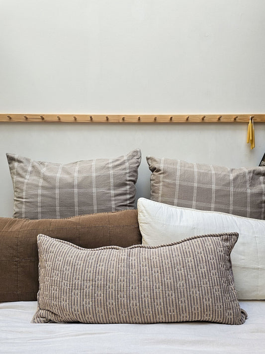 A collection of textured throw pillows in neutral tones, including a brown plaid pillow, a white patterned lumbar pillow, and a solid brown pillow, arranged on a bed against a light gray wall with a wooden peg rail.