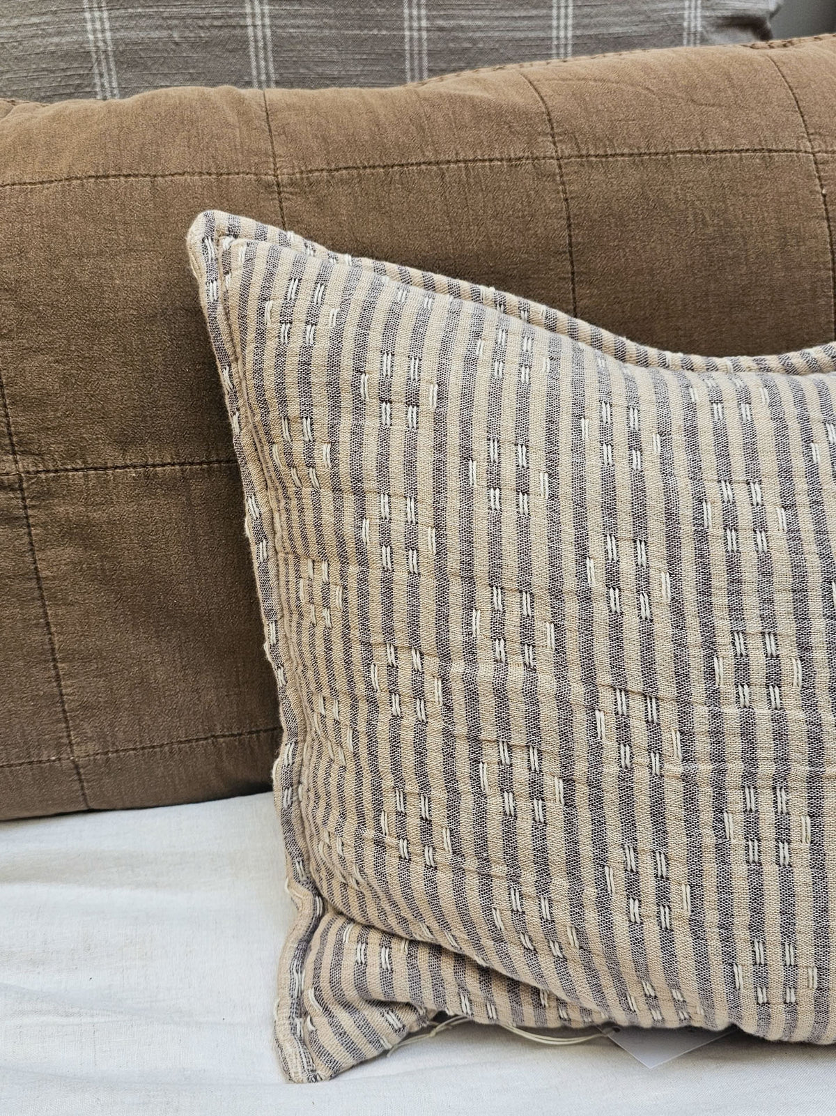 A square throw pillow with a beige and gray striped textured pattern, styled on a white surface in front of a brown quilted sofa cushion.
