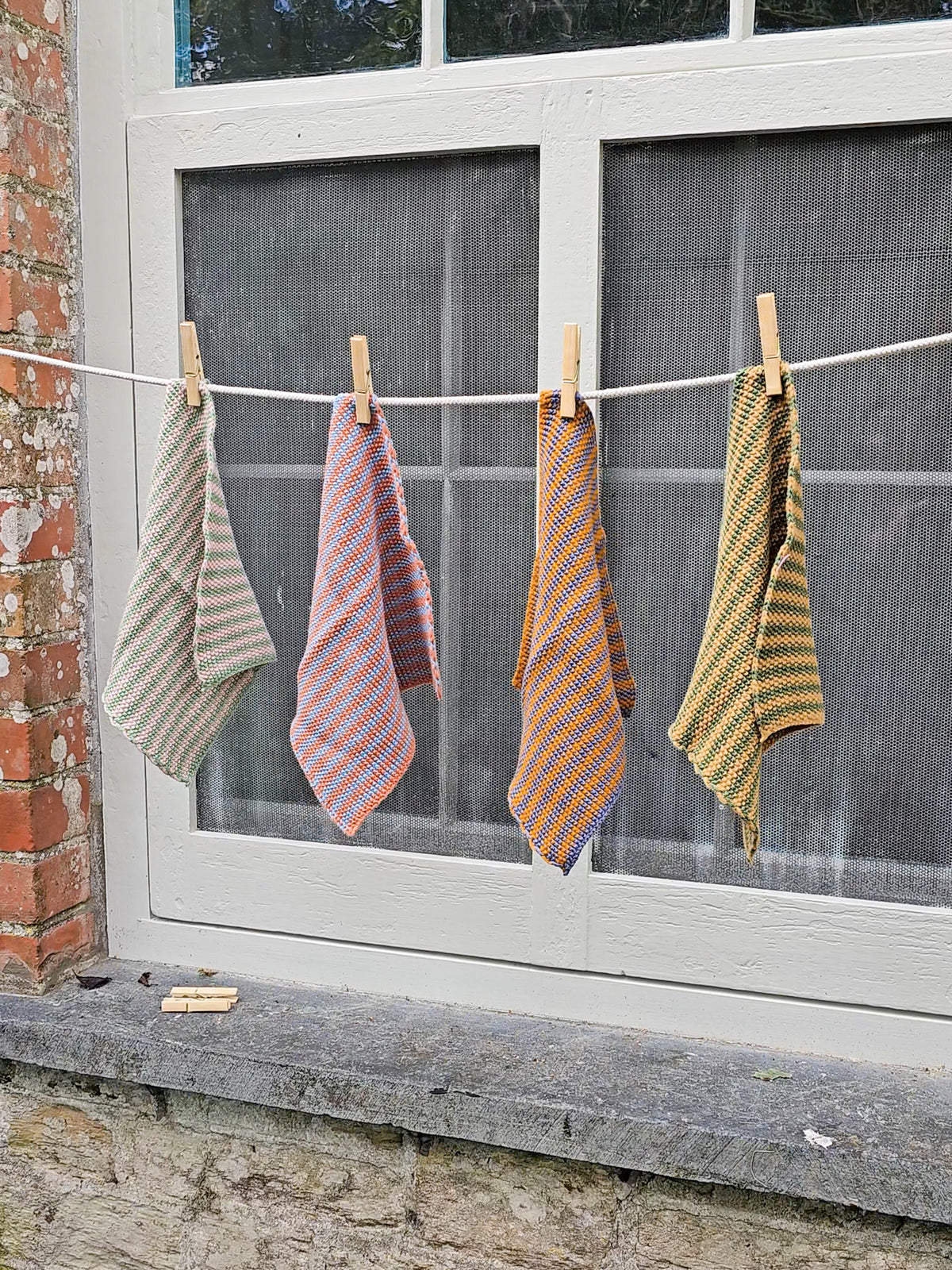 Four patterned kitchen dish cloths hanging from a white clothesline with wooden clothespins in front of a window.