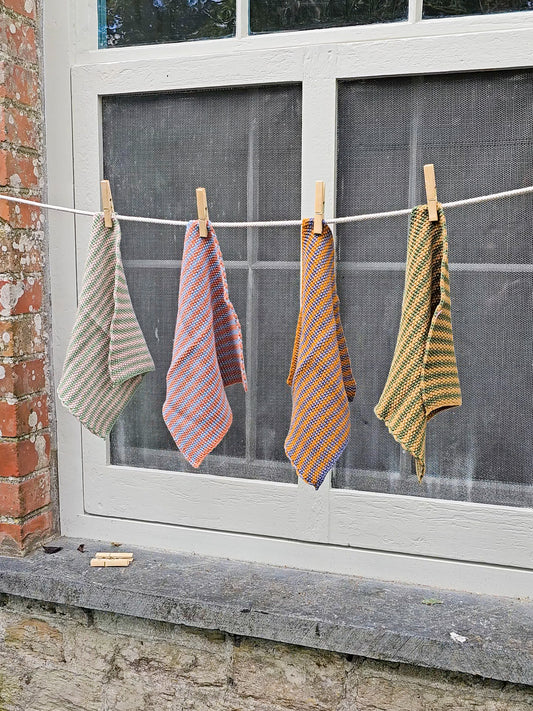 Four patterned kitchen dish cloths hanging from a white clothesline with wooden clothespins in front of a window.