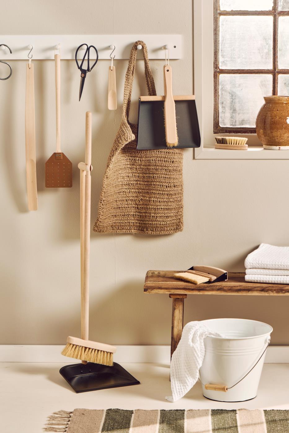 Cleaning supplies including a broom, dustpan, and bucket on a hook rack against a neutral wall.