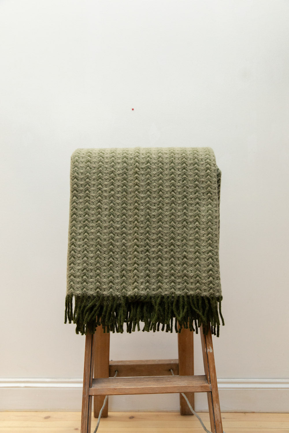 Ilse Wood wool blanket in sage green with chevron weave texture and dark green knotted fringe draped over a wooden stepladder.
