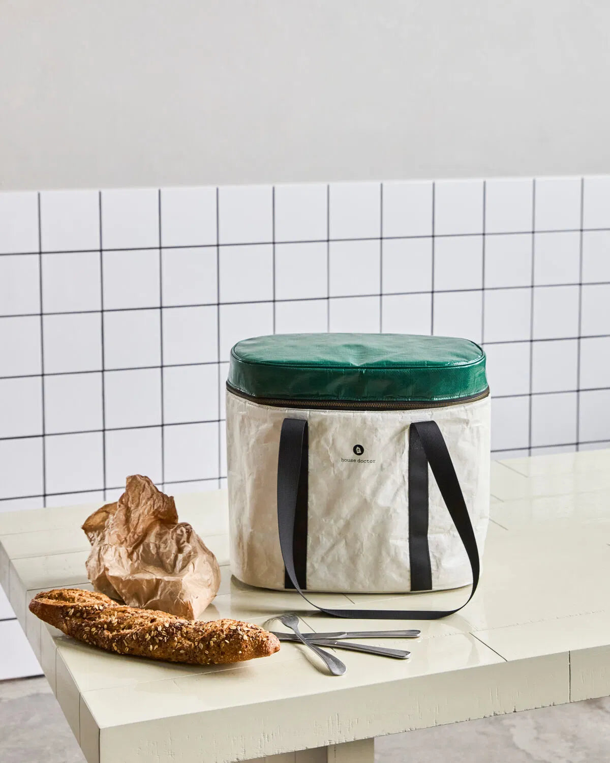 Insulated sand and green cooling picnic bag, shown in a kitchen setting, sat on top of a table, against a tiled wall. A seeded bread stick and cutlery sits next to it.
