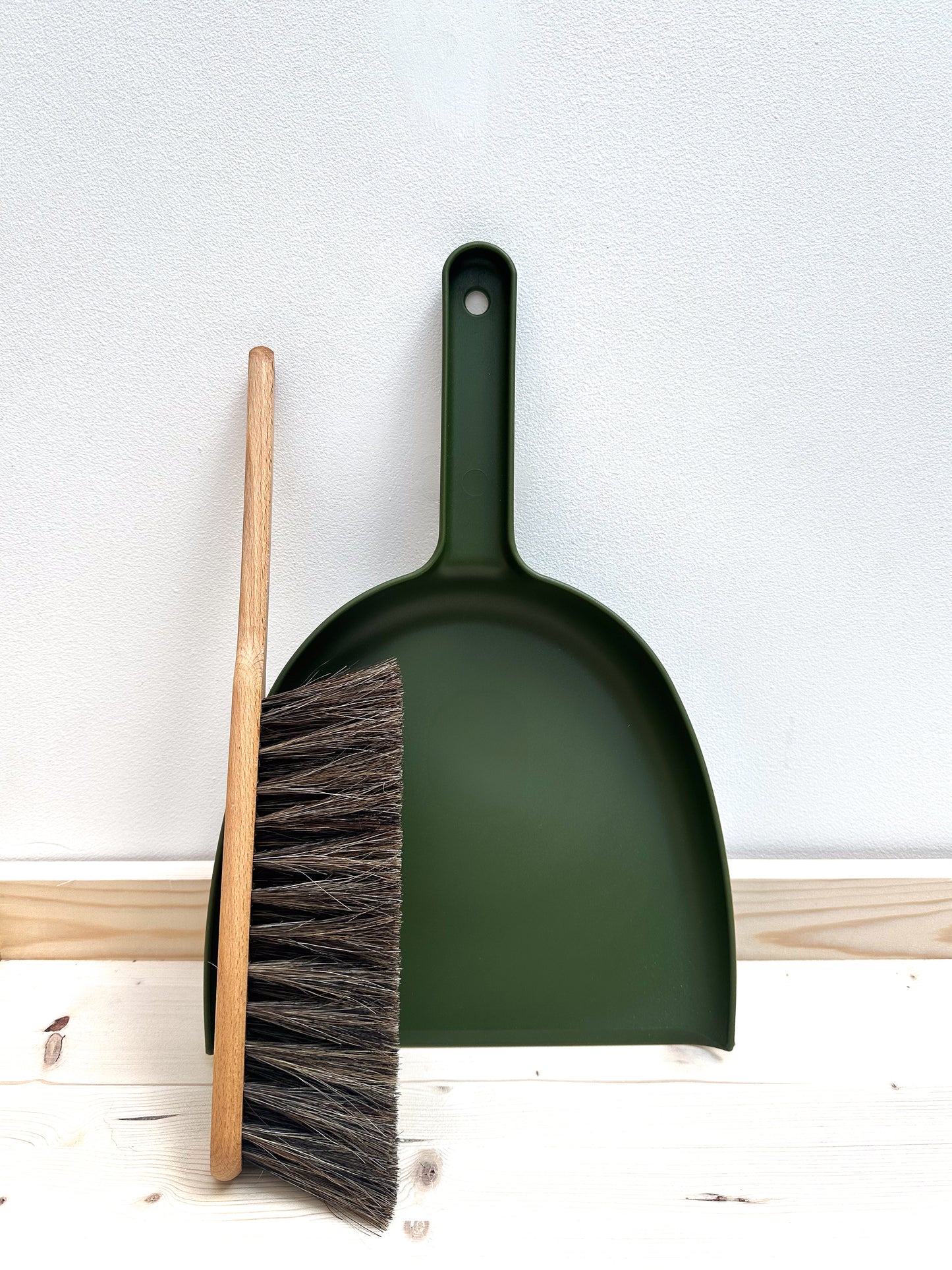 A moss green dustpan and a wooden brush with horsehair, placed against a white background.