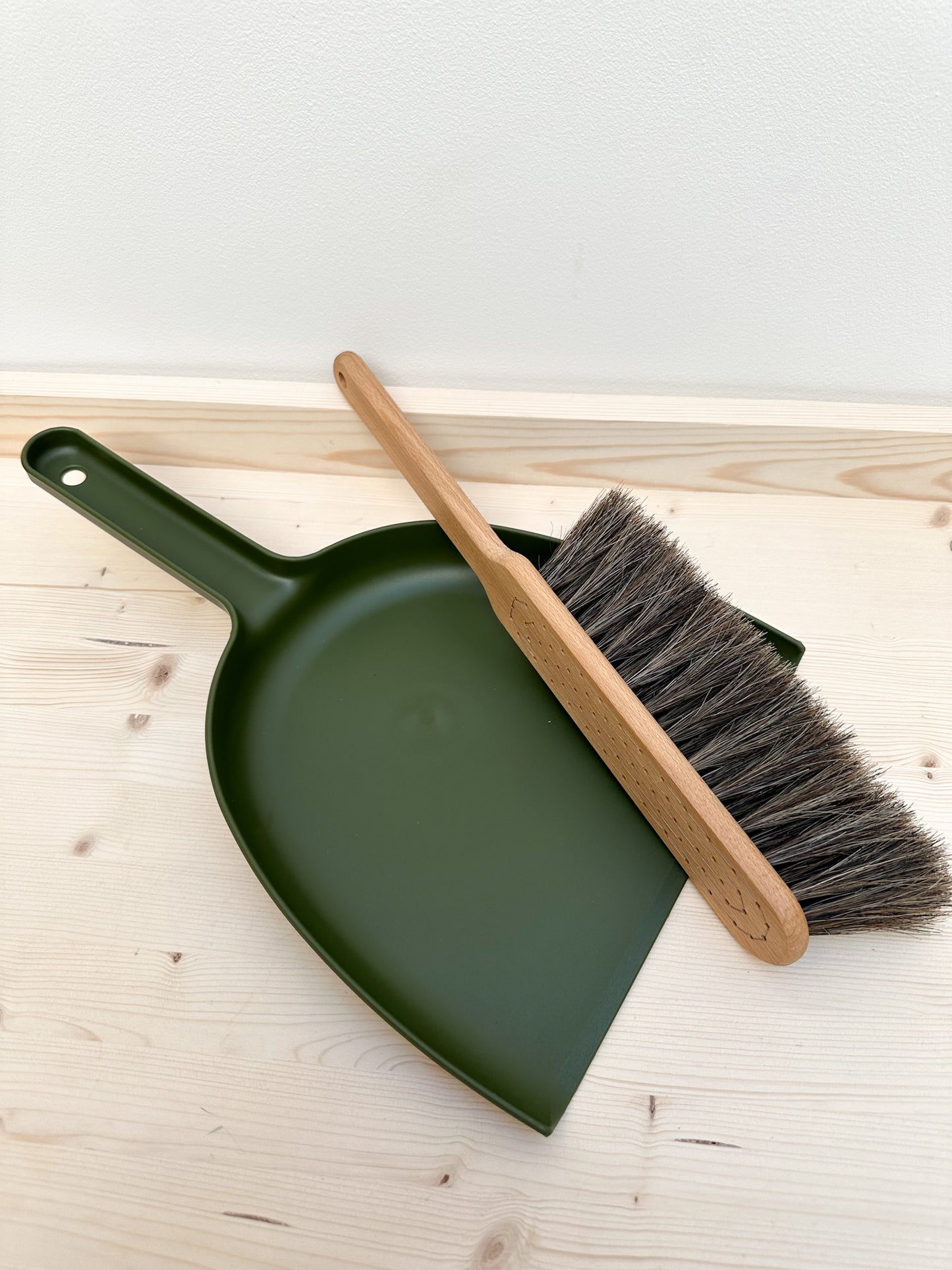 A moss green dustpan and wooden brush with horsehair bristle lays against a wooden surface.
