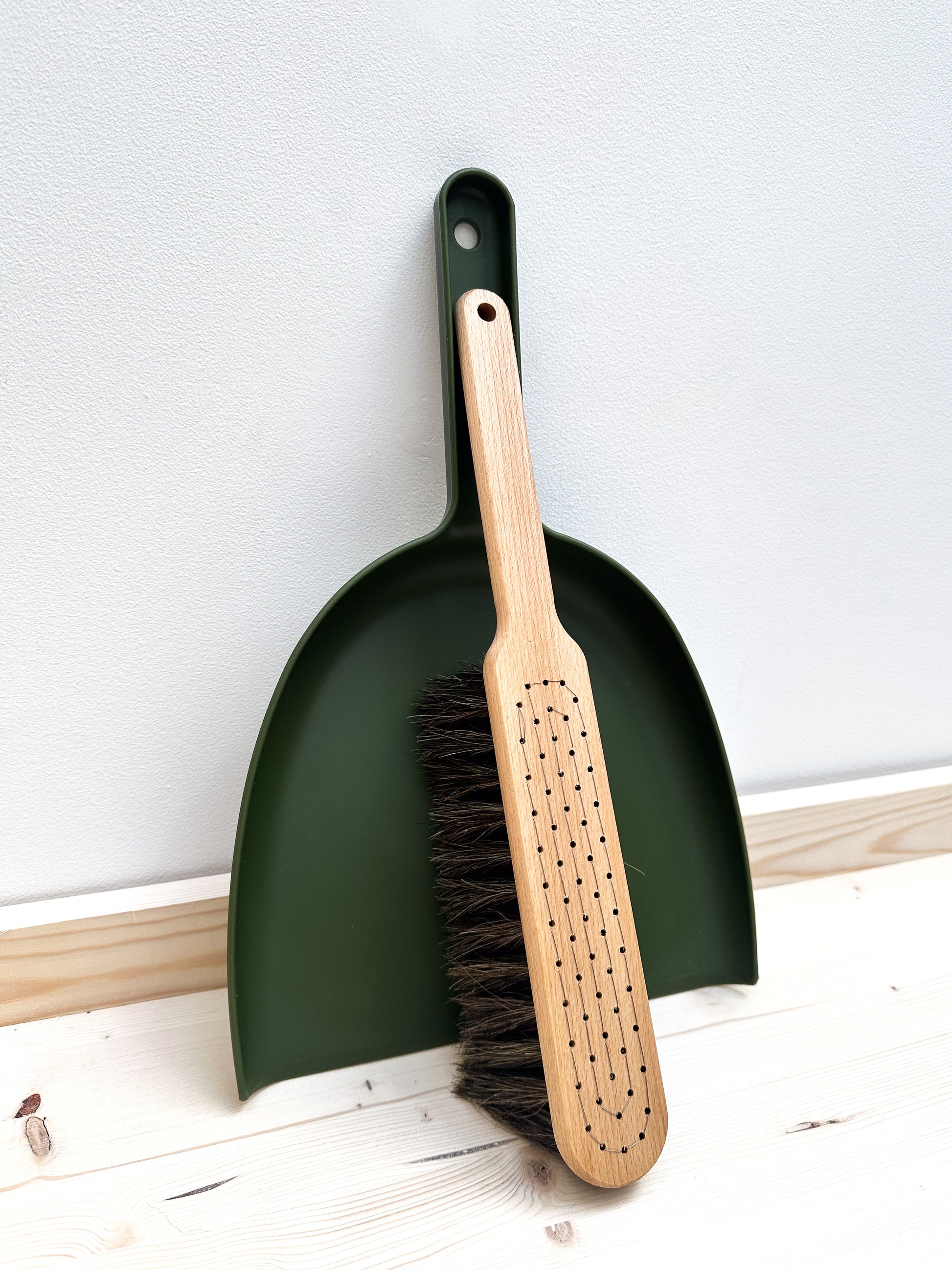 A moss green dustpan and wooden brush with horsehair bristle sits against a white wall on a wooden surface.