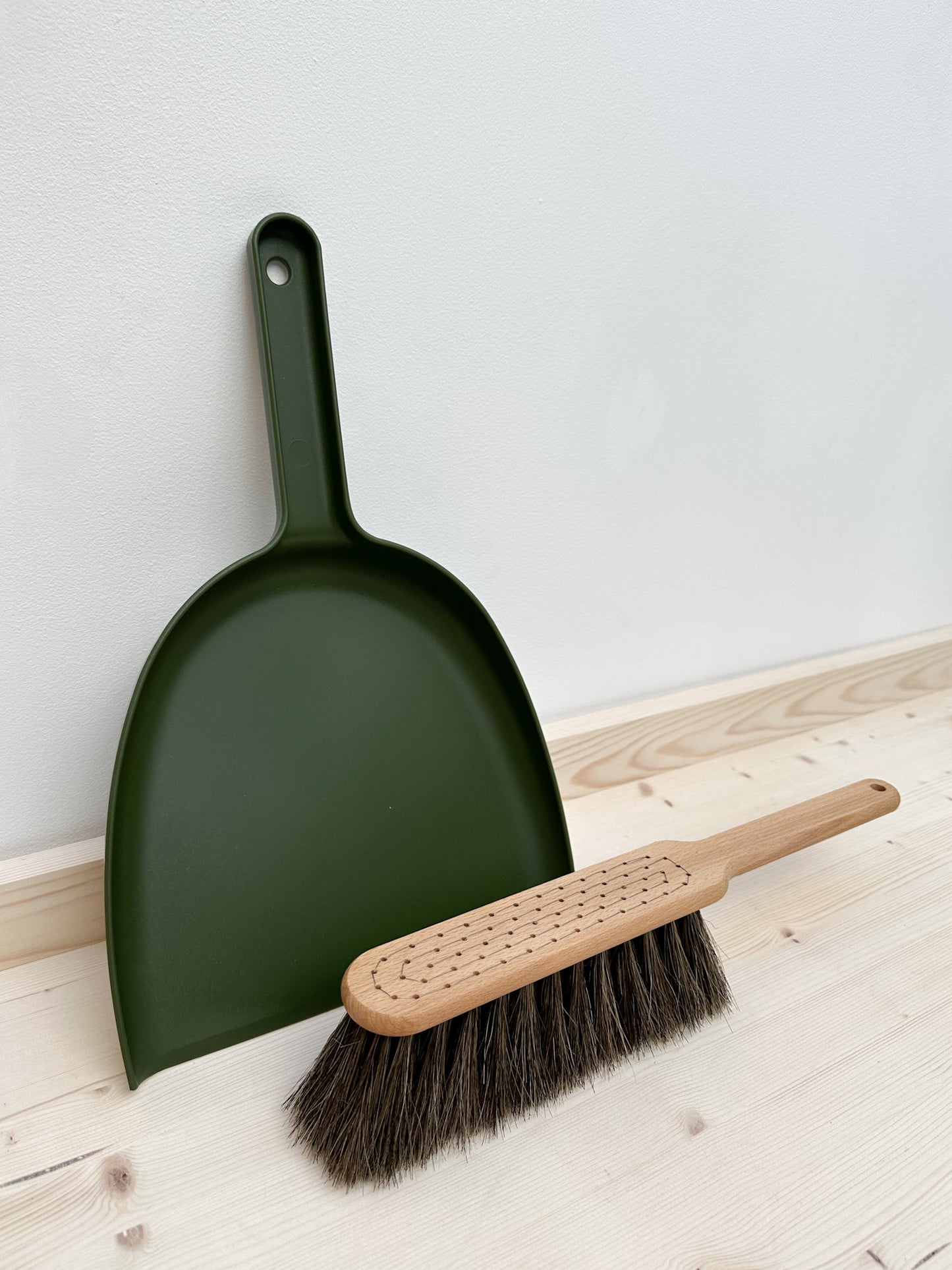 A moss green dustpan and wooden brush with horsehair bristle sits against a white wall on a wooden surface.