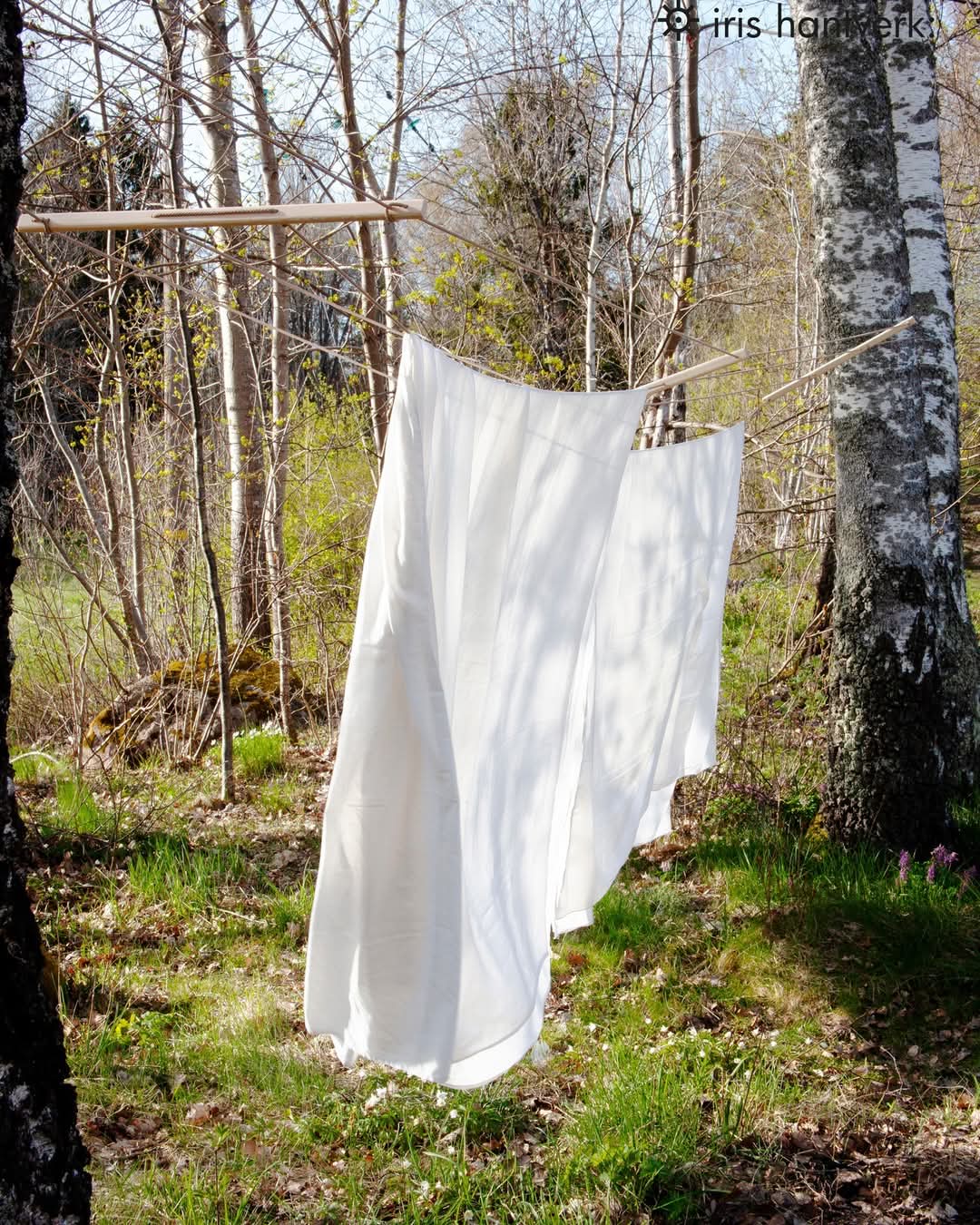 A clothesline made of pine and rope, with white fabric hanging on it, set against a backdrop of a garden with trees.