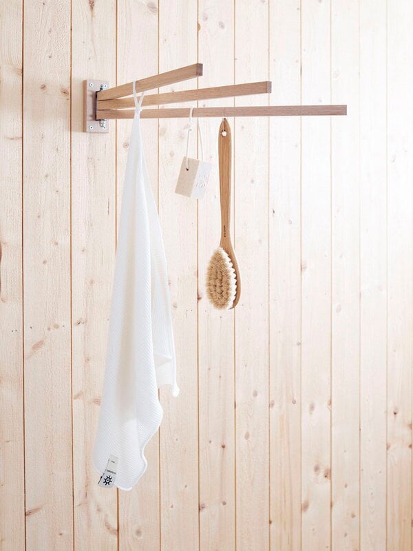 A wall-mounted towel dryer made of oiled oak, with towels and a brush hanging on it against a wooden backdrop.