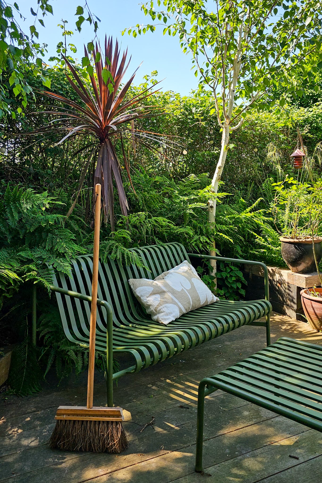 Iris Hantverk long handle wooden broom with natural bristles leaning against a green garden sofa on a decked terrace