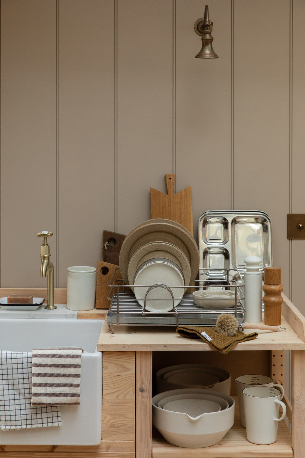 Pale wood kitchen with a butler sink, dish rack of plates, chopping boards and a brass tap