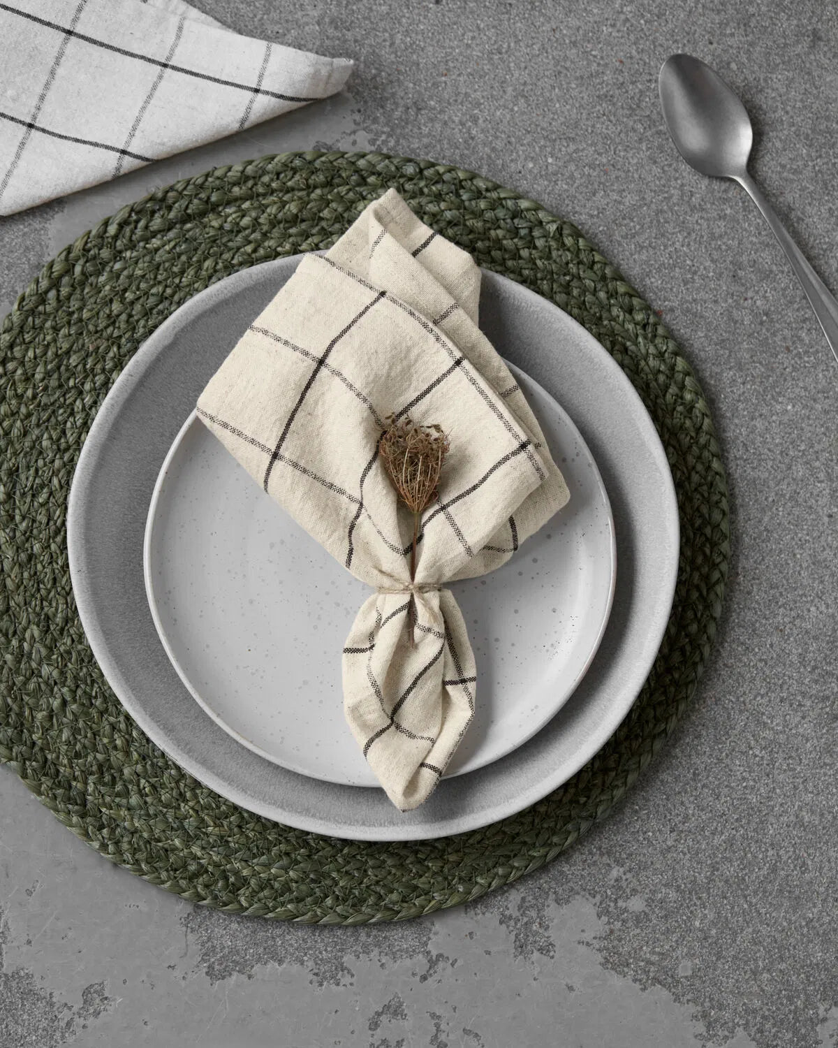 A white and black checked napkin is laying on two white plates stacked on top of each other on top of a green placemat, on a concrete table. A spoon lays nearby as well as the corner of another napkin.