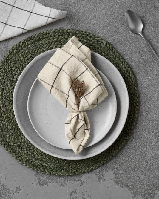 A white and black checked napkin is laying on two white plates stacked on top of each other on top of a green placemat, on a concrete table. A spoon lays nearby as well as the corner of another napkin.