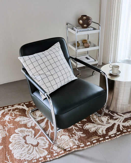A Join, off-white and black check cushion sits on a black leather and chrome chair. The chair is on a brown patterned rug, next to a chrome table, with a trolley in the background.