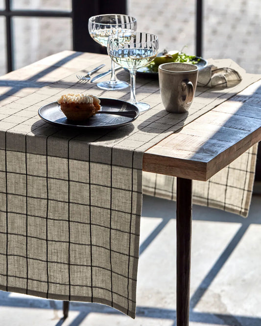 Off-white and black check Join table runner shown on a wooden table, with two glasses, two plates and a mug placed on top.