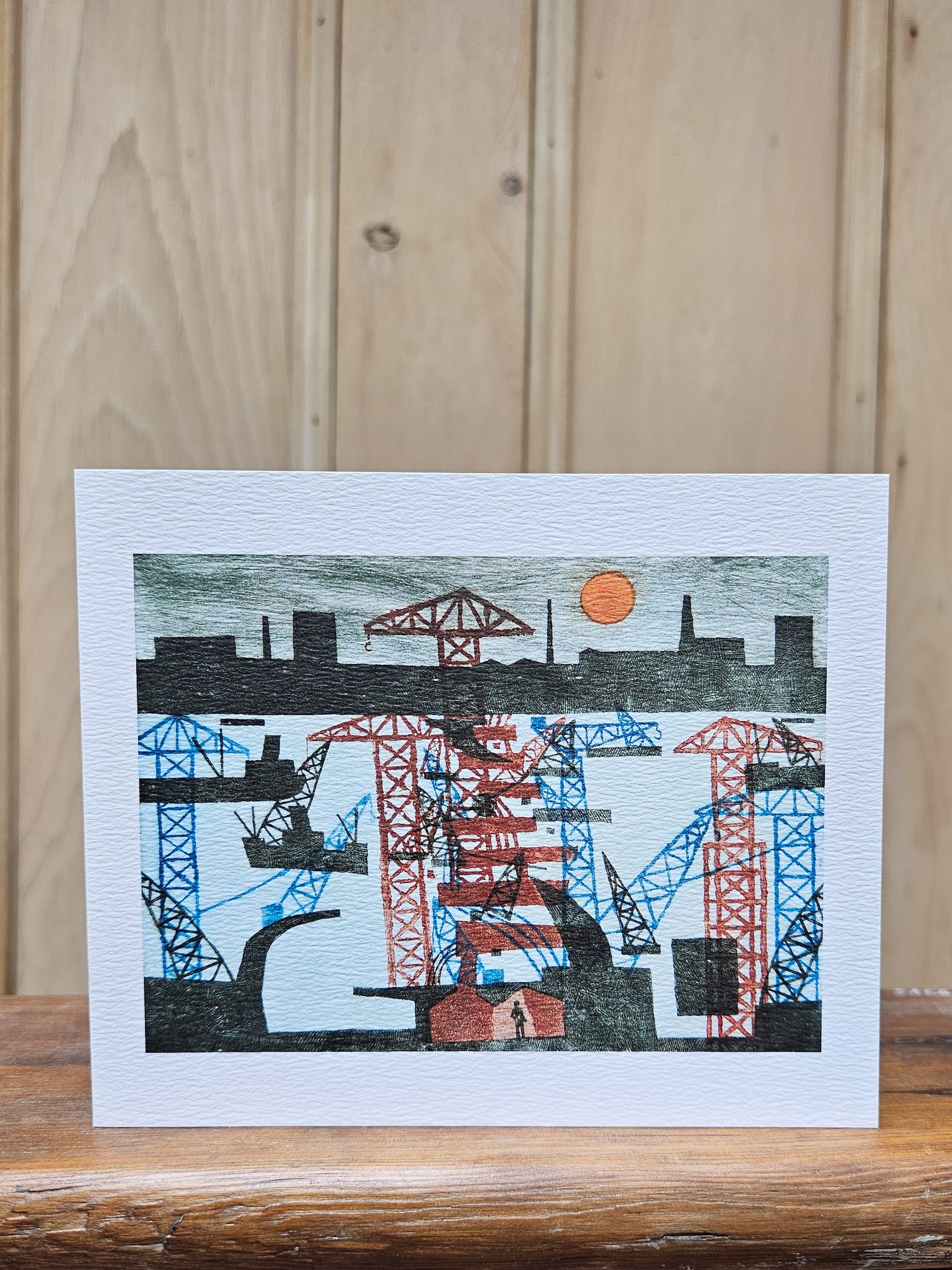 Greeting card reproducing Julian Trevelyan's print of the Woolwich Barrier construction, showing a tangle of red and blue cranes and industrial structures on the Thames beneath an orange sun
