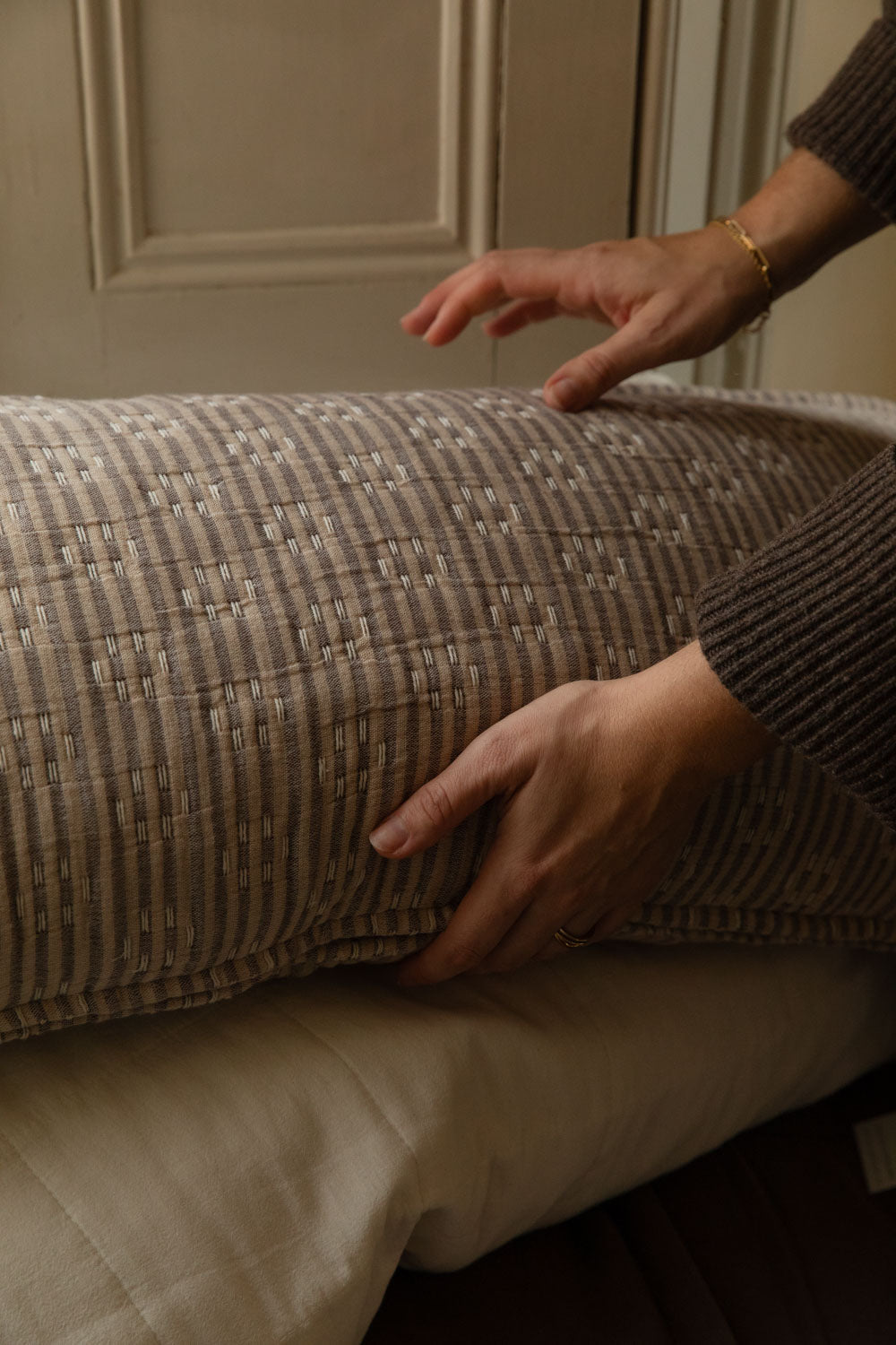Close-up of hands plumping a Kantha stitch striped cushion, showing the natural and brown hand-stitched texture