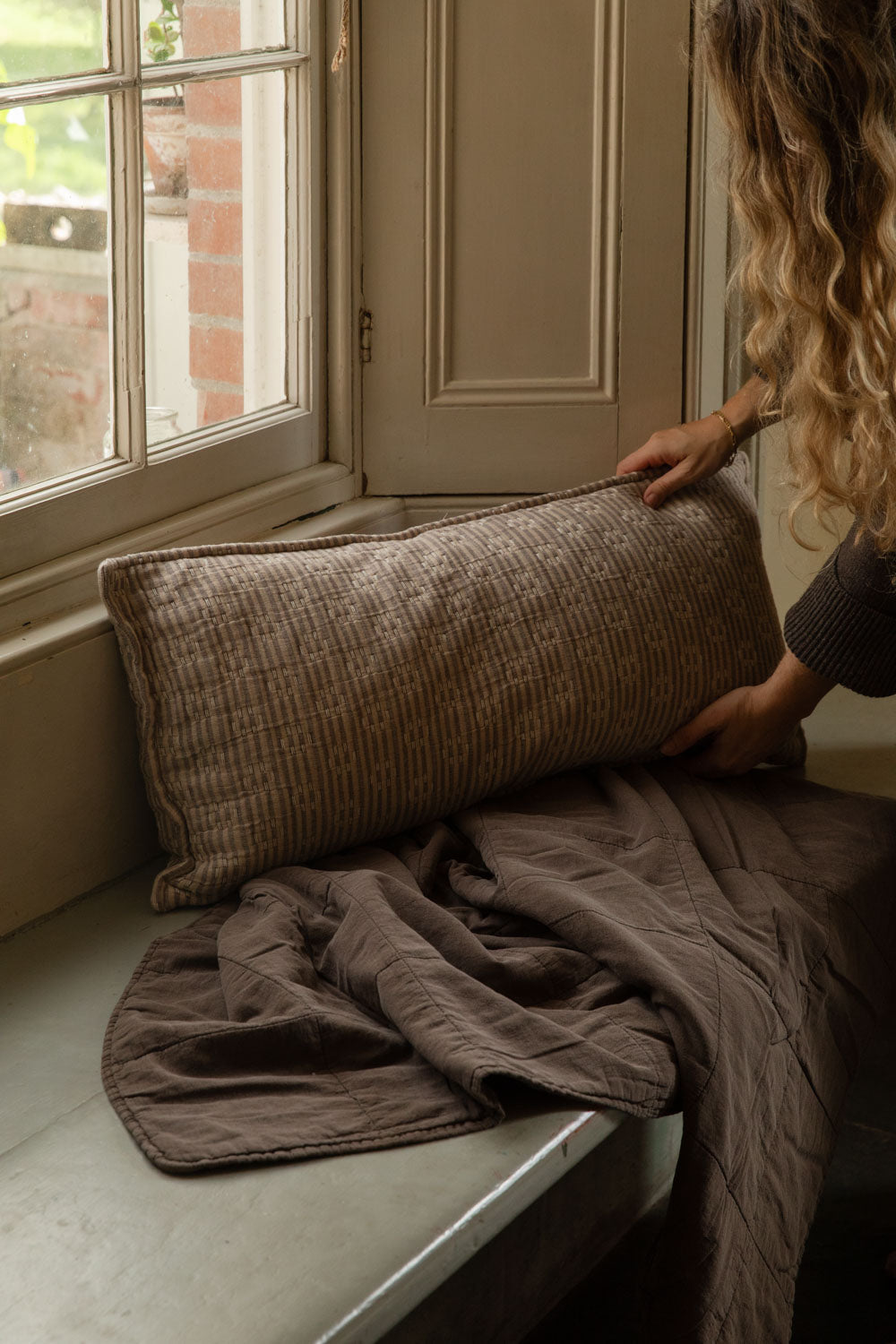 Hands placing a Kantha stitch striped cushion in natural and brown onto a window seat over a brown cotton quilt