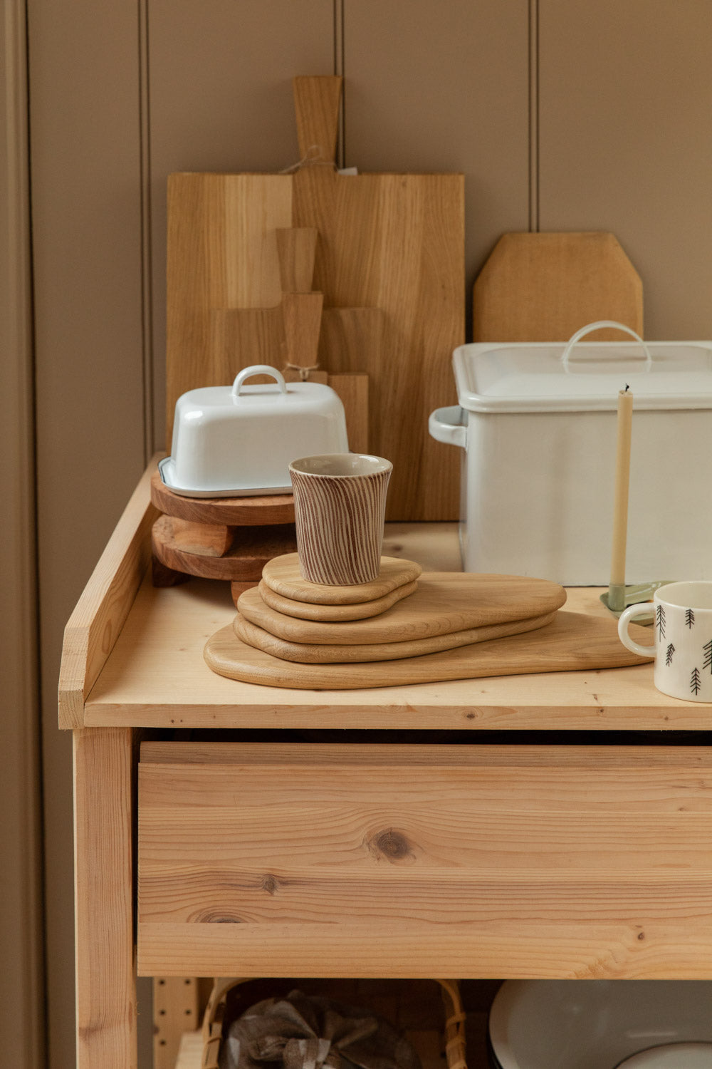 Pine kitchen table styled with stacked oak serving boards, Diva stripe mug, white enamel butter dish, enamel bread bin, and tree print mug against a neautral panelled wall.