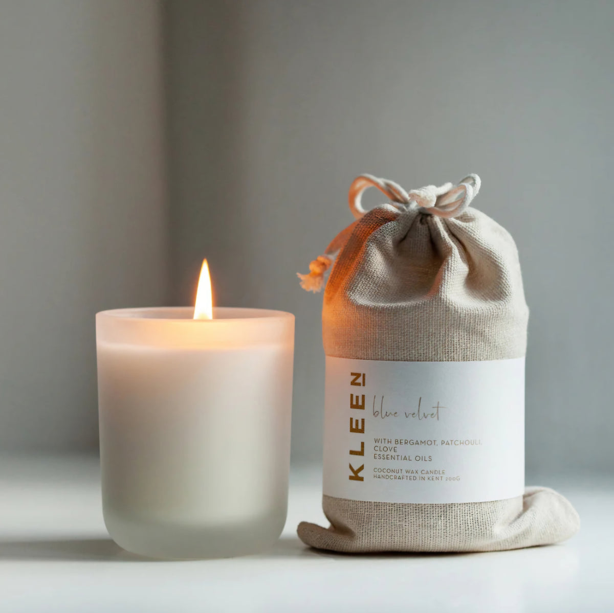 Kleensoaps Blue Velvet coconut wax candle lit beside its cotton drawstring bag, scented with bergamot, patchouli and clove