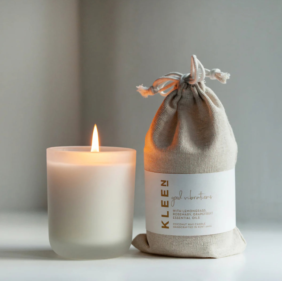Kleensoaps Good Vibrations coconut wax candle lit beside its cotton drawstring bag, scented with lemongrass, rosemary and grapefruit