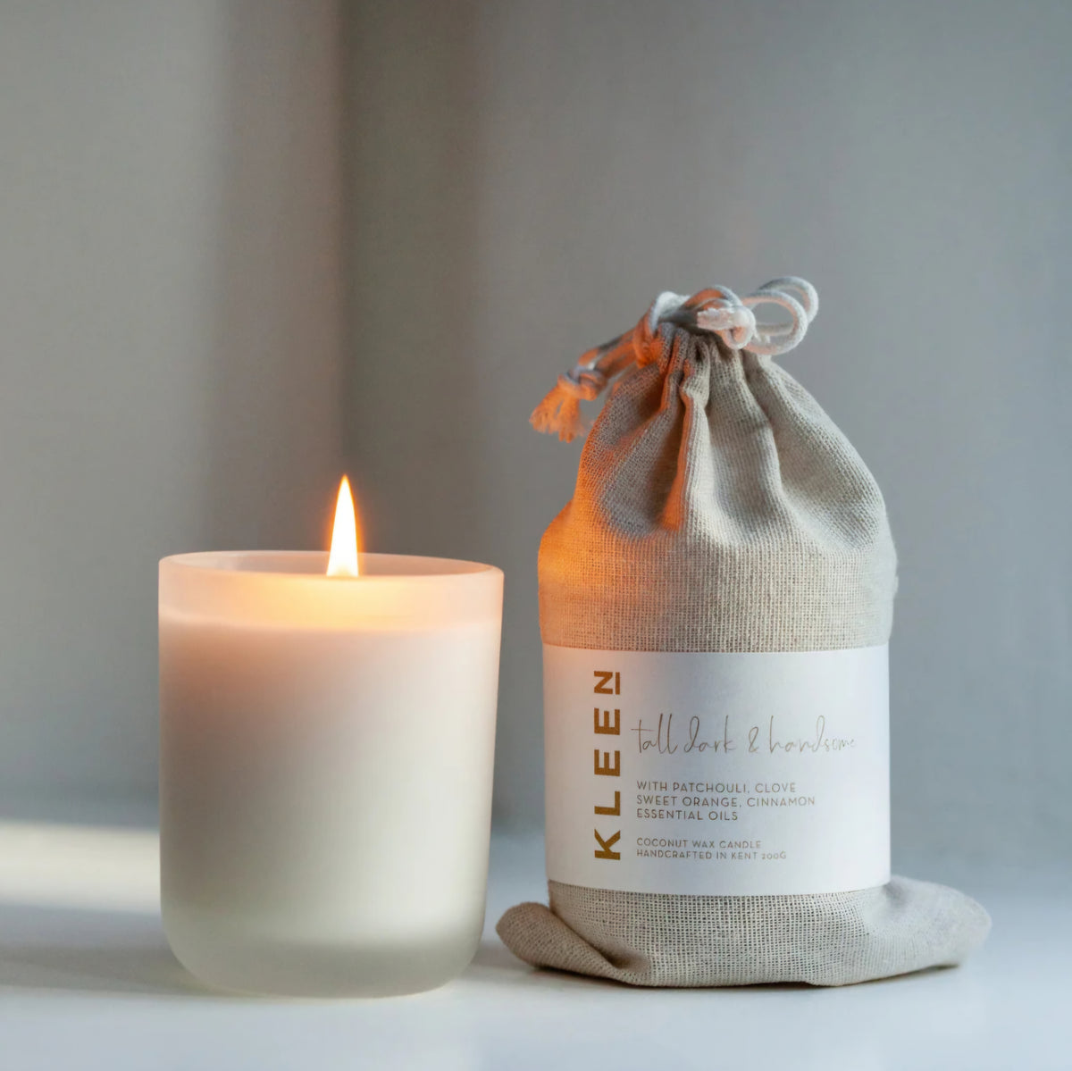 Kleensoaps Tall Dark and Handsome coconut wax candle lit beside its cotton drawstring bag, scented with patchouli, clove, sweet orange and cinnamon