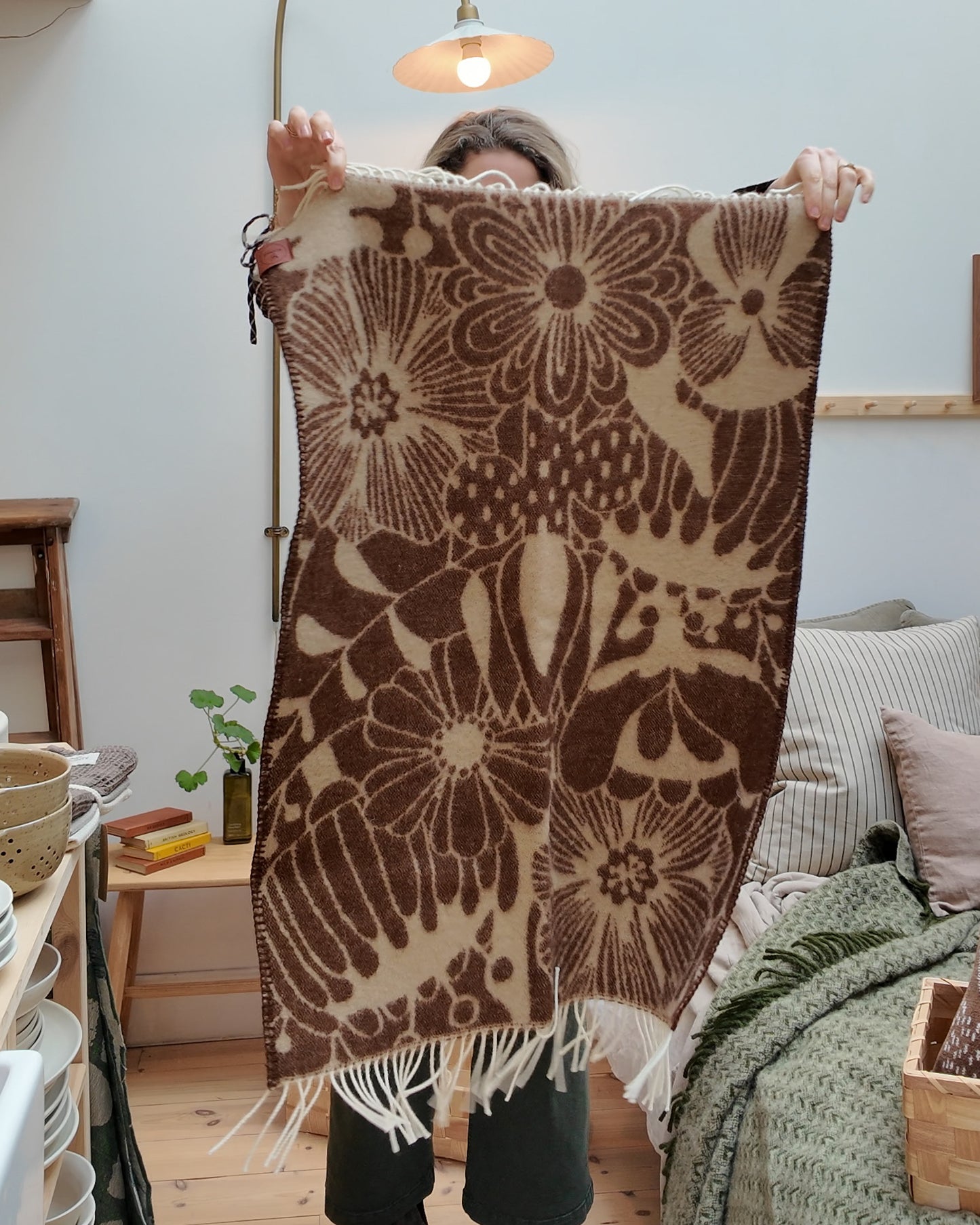 A woman holds up a Klippan Blomranka lambswool baby blanket in chocolate brown and sand with bold floral print and cream fringe edge.