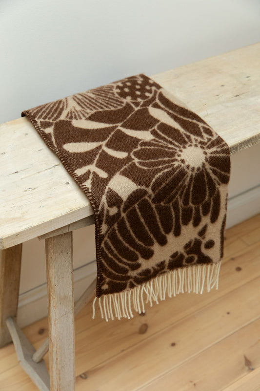 Klippan Blomranka lambswool baby blanket in chocolate brown and sand with bold floral print and cream fringe edge draped over a rustic pale wood table.