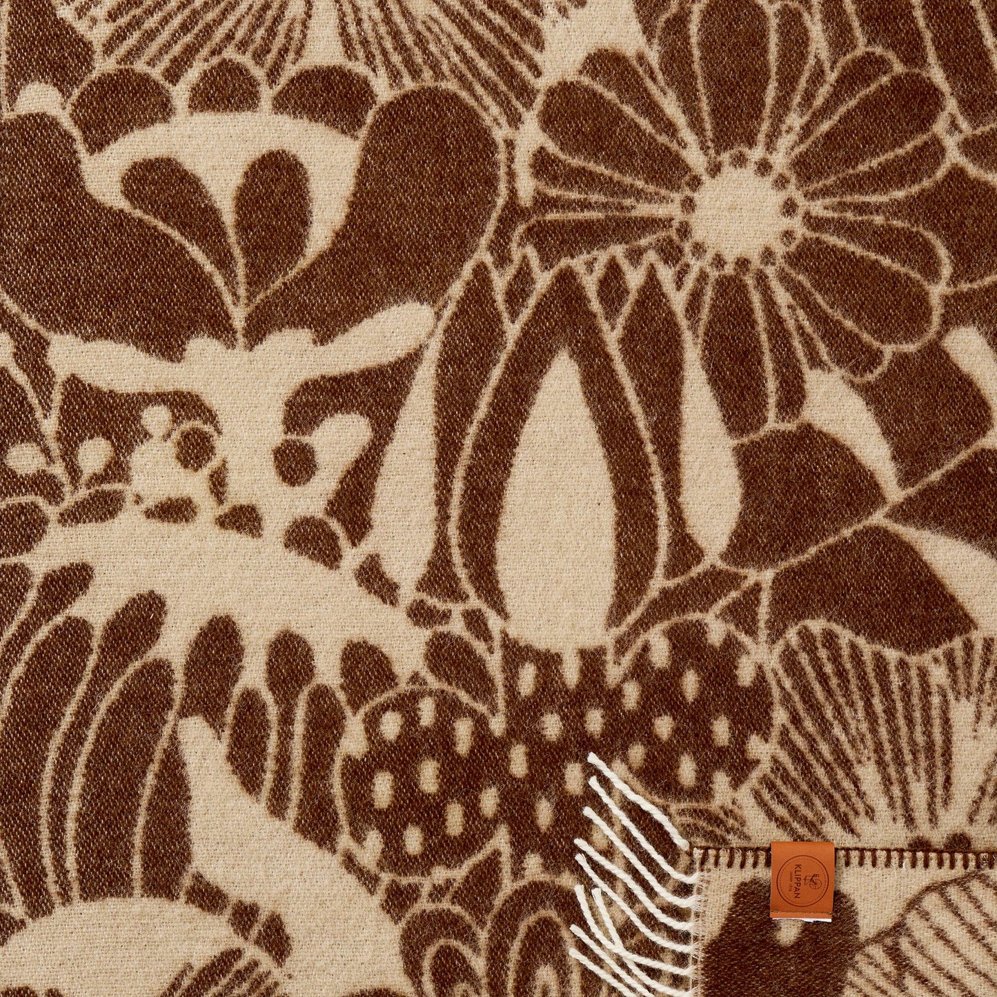 Close-up of Klippan Blomranka lambswool baby blanket in chocolate brown and sand with bold floral print and cream fringe edge.