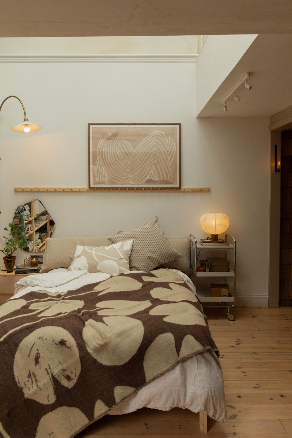 Scandinavian bedroom with Klippan Blomranka lambswool blanket in brown and cream draped over bed, Blomus Kami lamp on chrome trolley and framed artwork on peg rail.