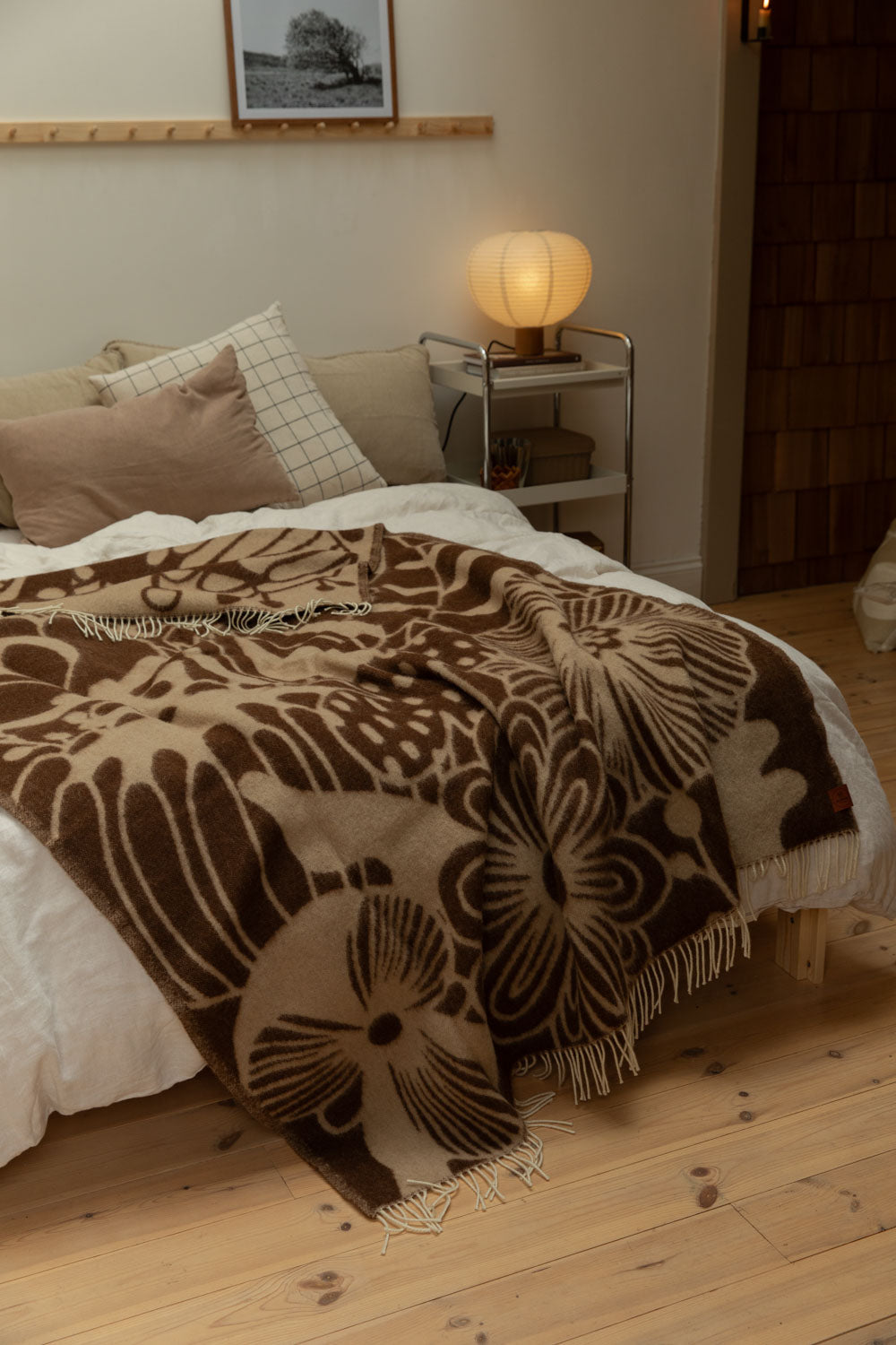 Klippan Blomranka lambswool blanket in chocolate brown and sand with bold floral print draped generously over a bed with chrome trolley bedside table, glowing paper lantern lamp and pine floorboards.