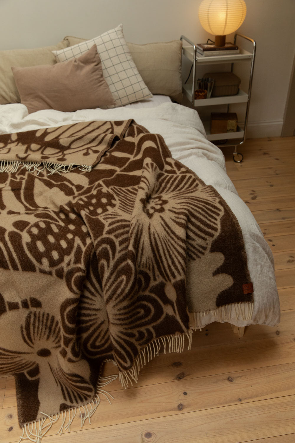 Klippan Blomranka lambswool blanket in chocolate brown and sand draped generously over a bed with chrome trolley bedside table and glowing paper lantern lamp.